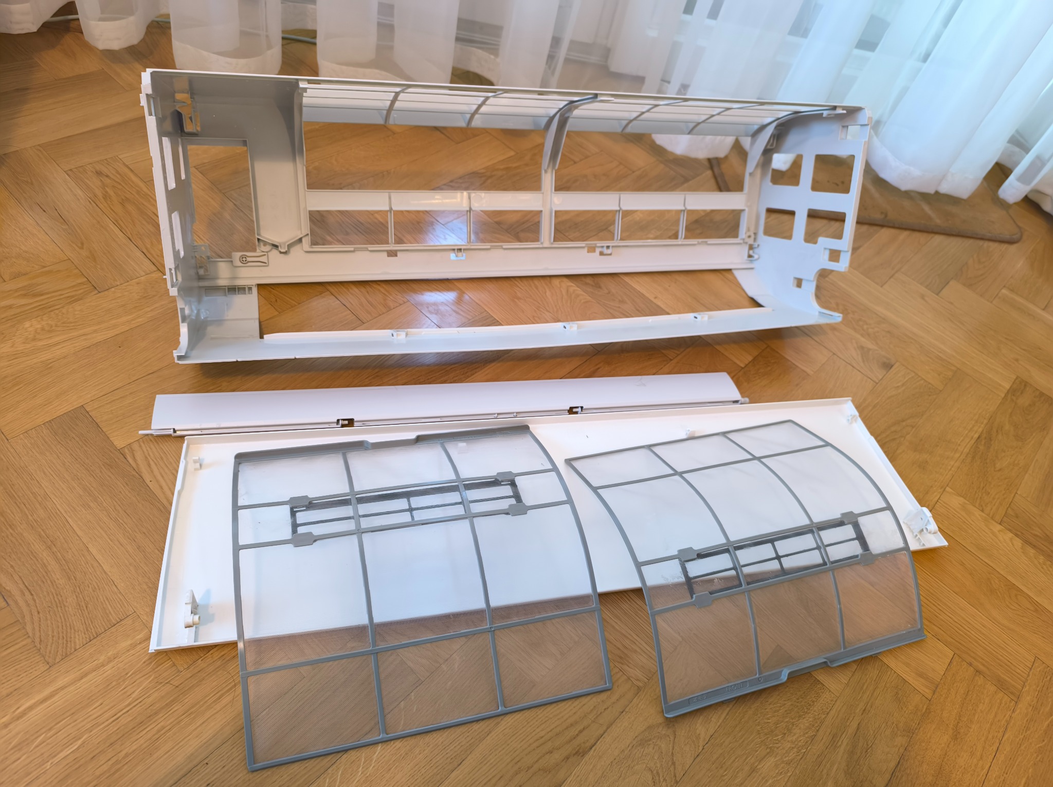 Rozłożony panel klimatyzatora z filtrami na parkiecie, gotowy do czyszczenia lub serwisu. Widoczne elementy obudowy i filtry powietrza.