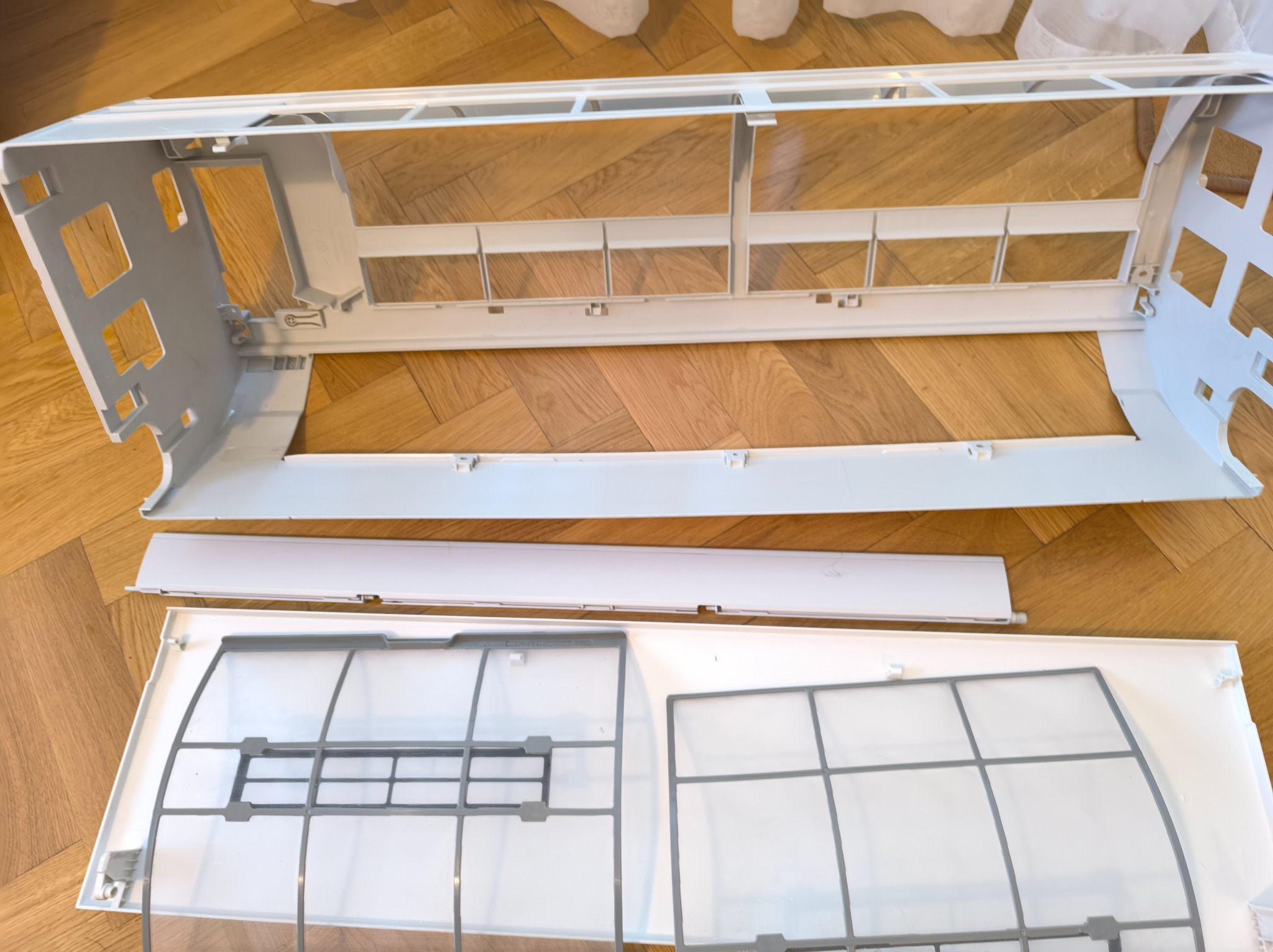 Rozłożony na parkiecie biały panel klimatyzacji z filtrami, ukazujący etapy czyszczenia lub konserwacji. Widoczne elementy składowe jednostki wewnętrznej.