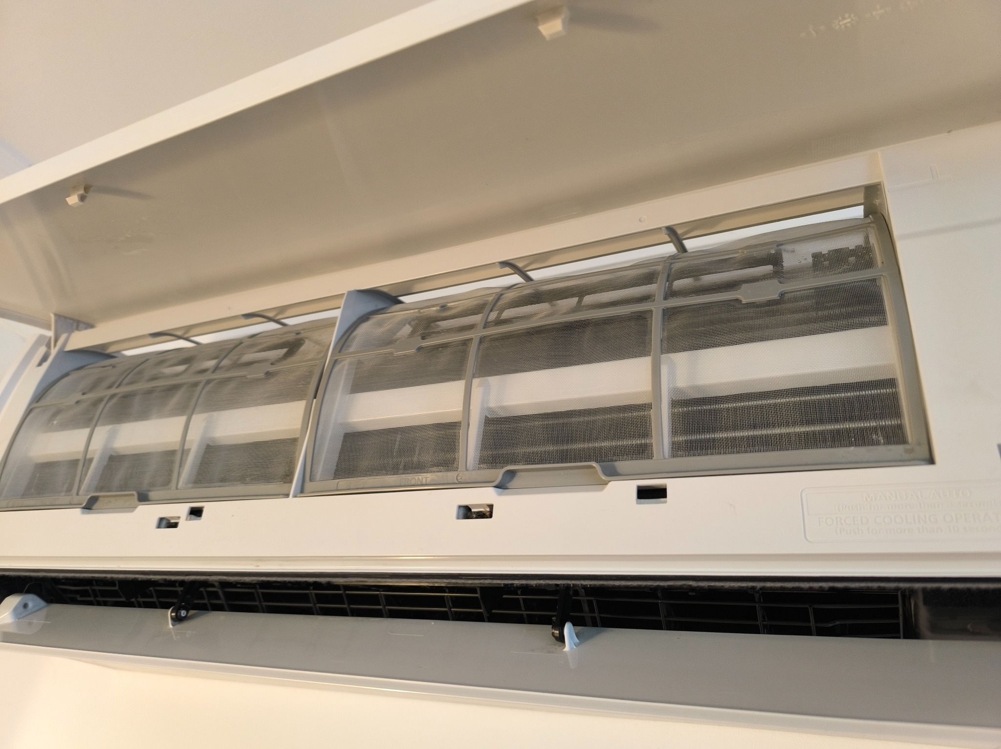 Otwarty, biały klimatyzator z widocznymi, zakurzonymi filtrami siatkowymi. Widoczne żebra chłodzące i obudowa z napisem 'Manual/Auto'.