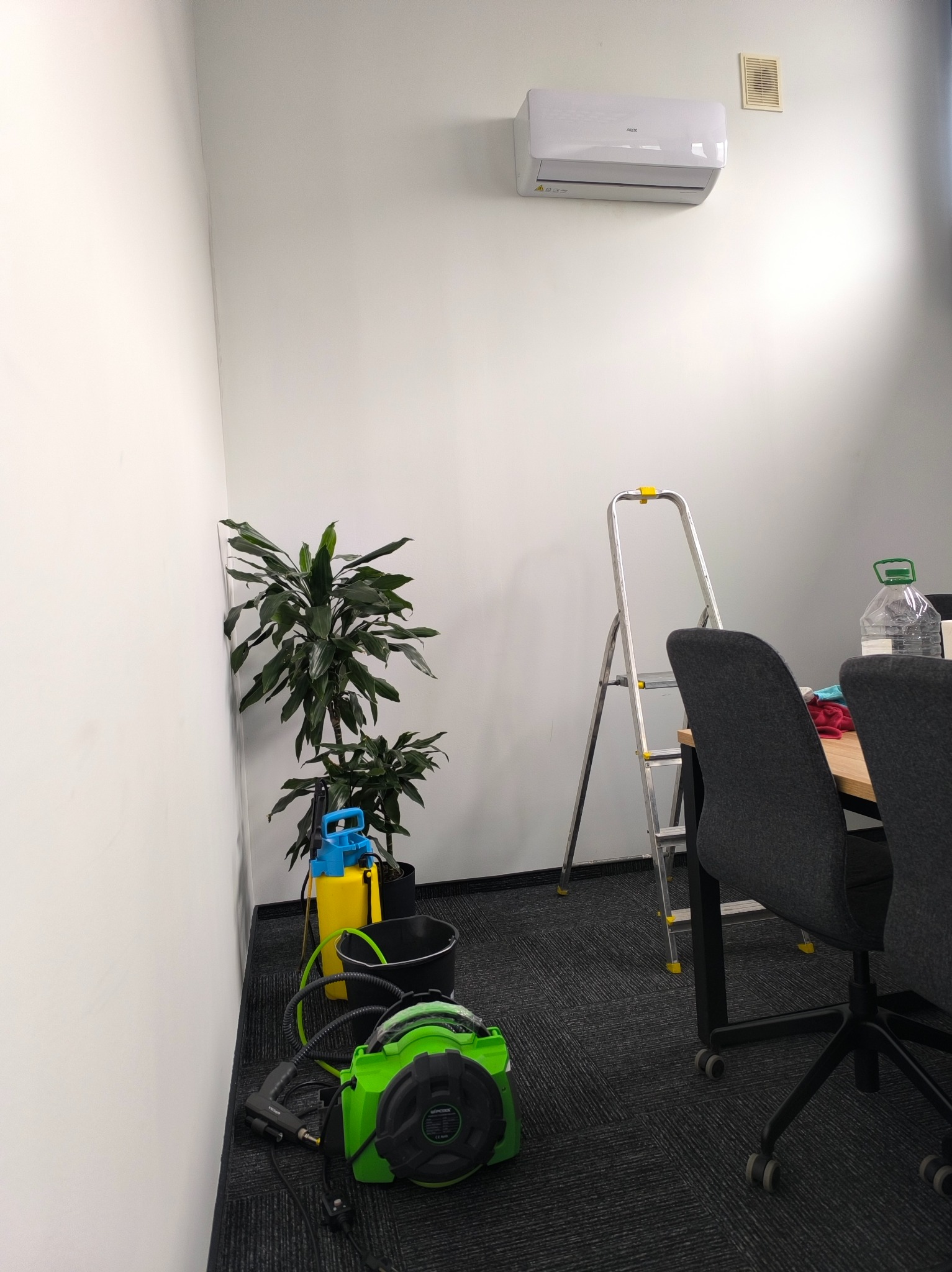 Biała klimatyzacja AUX na ścianie biura. Obok drabina, kwiaty i sprzęt czyszczący. Usługi montażu i serwisu w Strykowie.