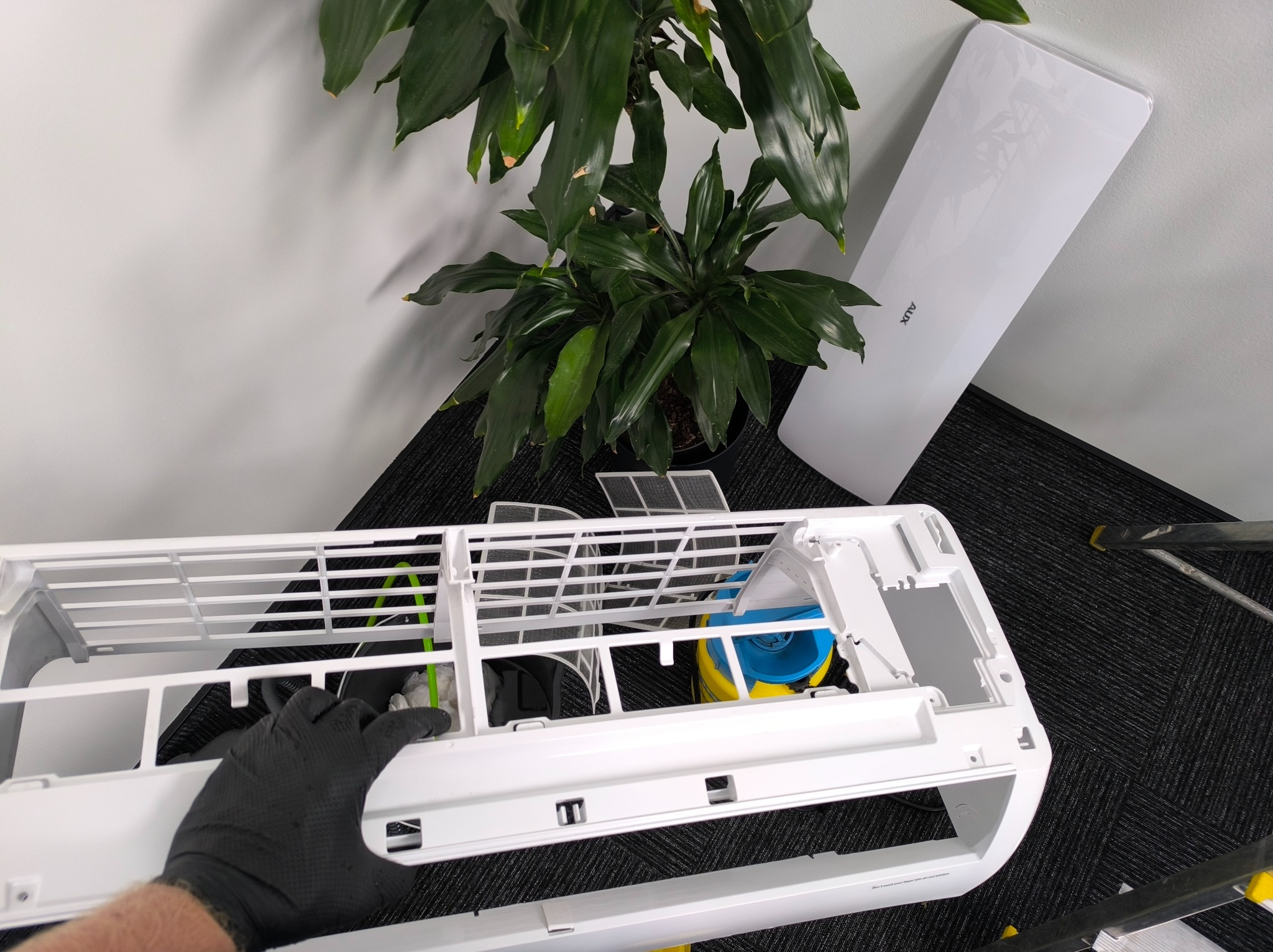Demontaż klimatyzacji AUX: widoczna obudowa, filtr i dłoń w czarnej rękawicy. W tle roślina doniczkowa i obudowa. Perspektywa z góry.