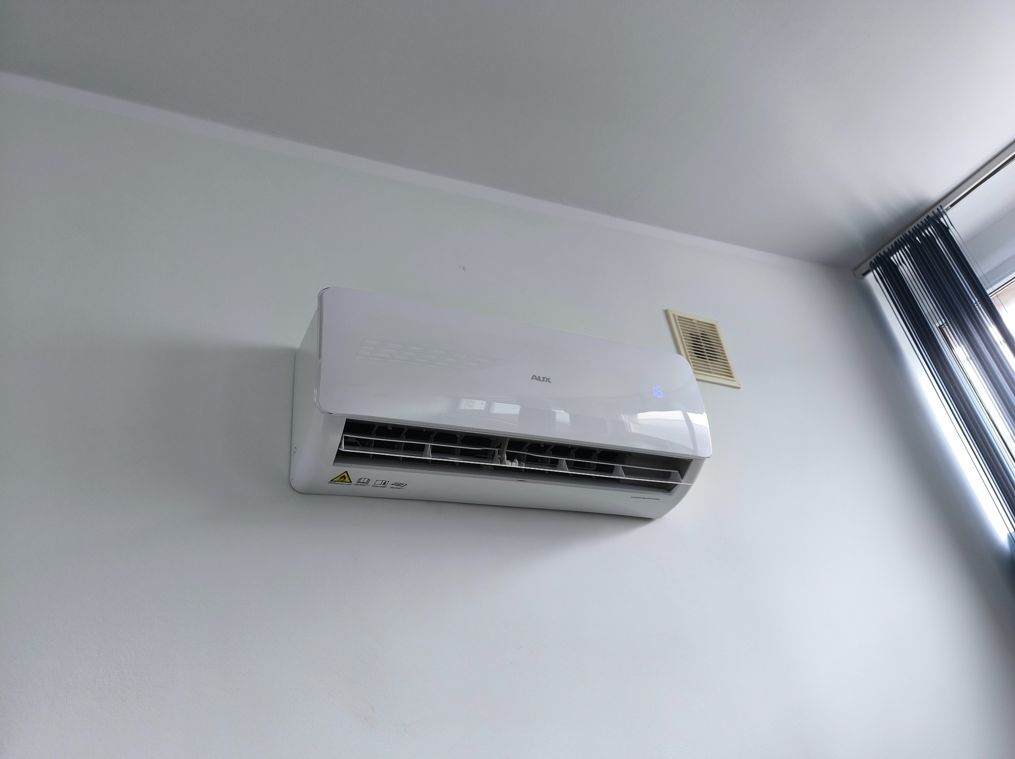 Biała klimatyzacja AUX zamontowana na ścianie w jasnym pomieszczeniu. Widoczna kratka wentylacyjna i fragment okna z zasłonami. Minimalistyczny design.