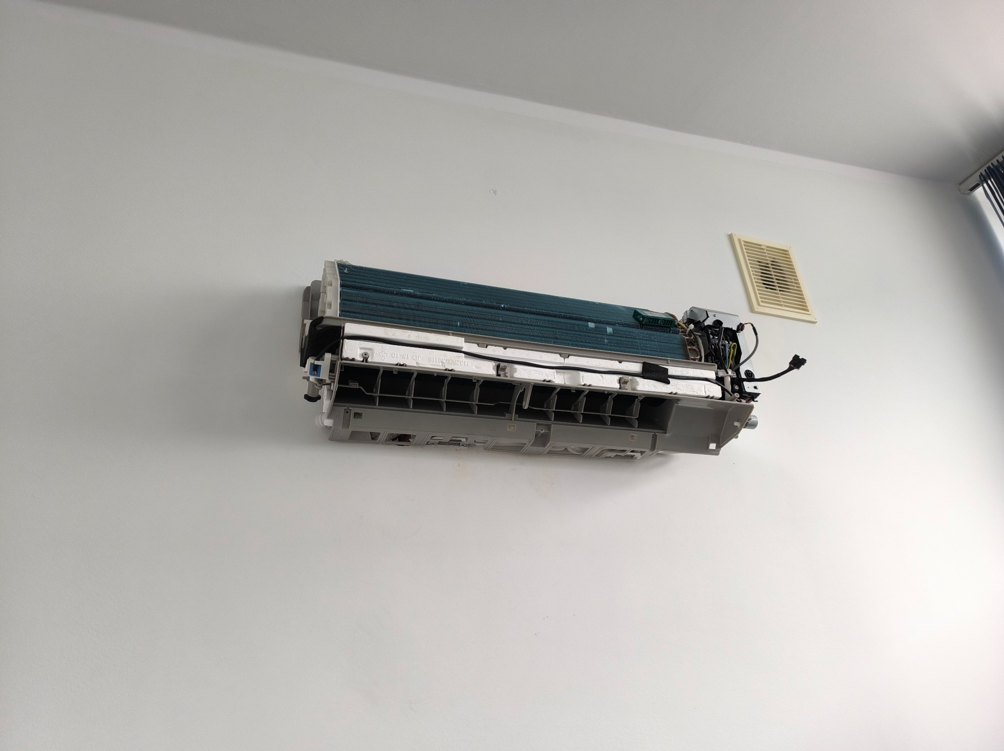 Otwarty klimatyzator ścienny, widoczne elementy wewnętrzne, na białej ścianie. Obok kratka wentylacyjna i fragment okna z zasłoną. Widok z dołu.