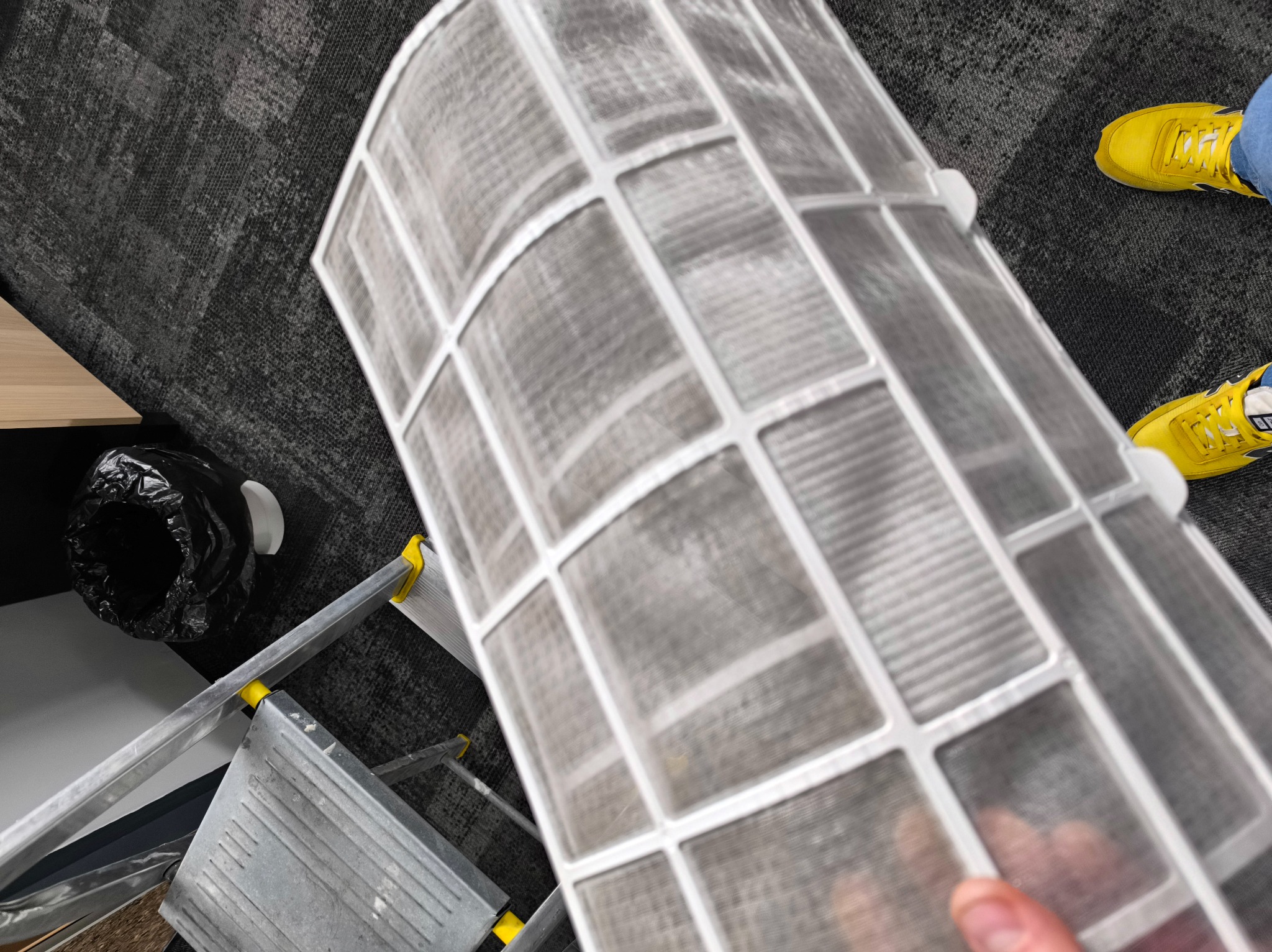 Brudny filtr klimatyzacji trzymany w dłoni, widoczna drabina, śmieci i żółte buty. Filtr wymaga czyszczenia lub wymiany.