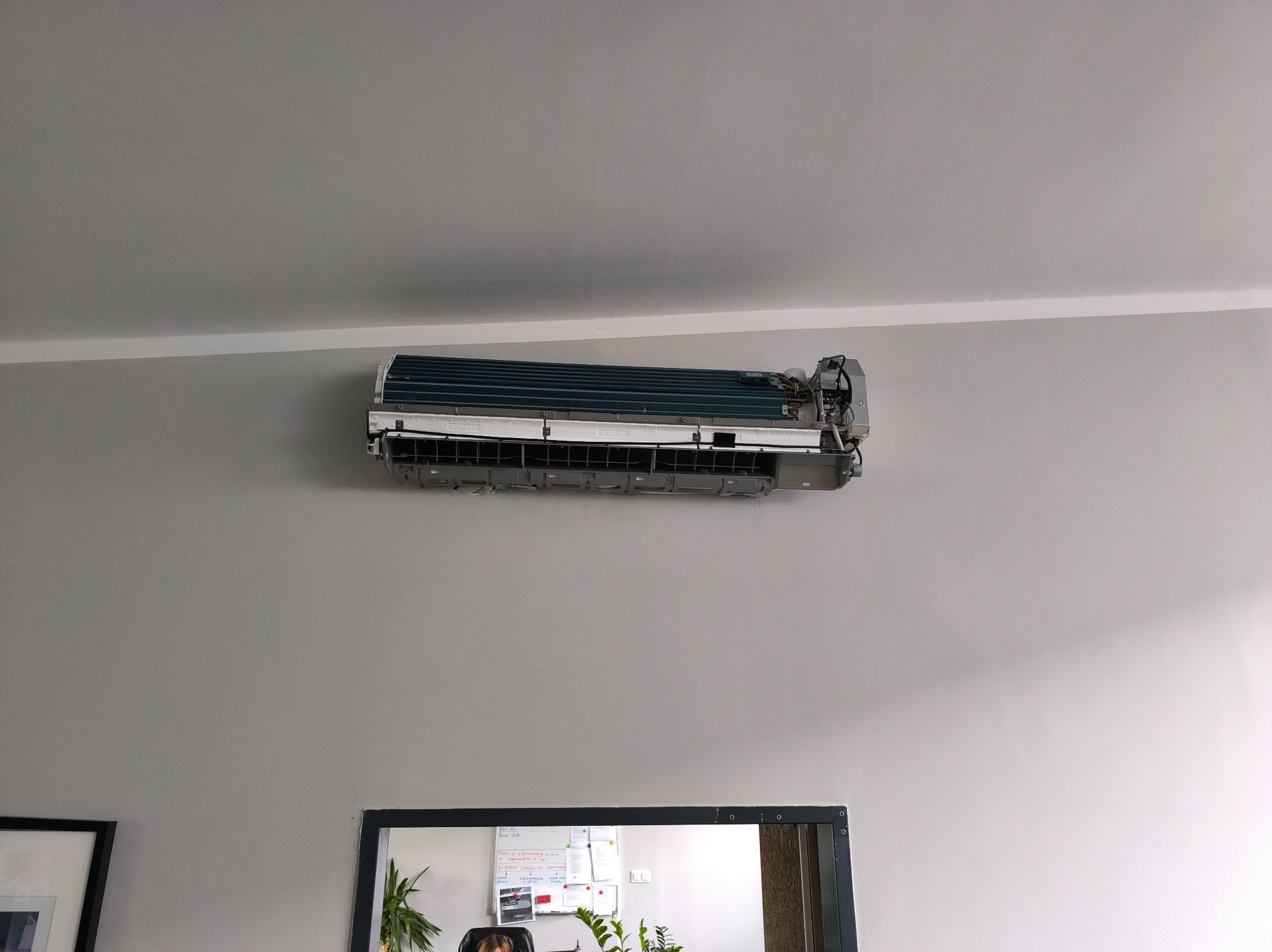 Rozmontowany klimatyzator na ścianie w szarym pomieszczeniu biurowym, widoczne elementy instalacji i chłodnica. Urządzenie w trakcie czyszczenia lub naprawy.