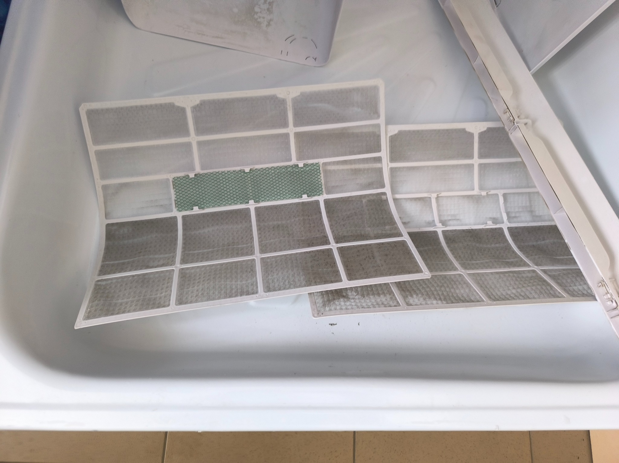 Dwa brudne filtry klimatyzacji w białej wannie, jeden z zielonym elementem filtrującym. Widoczne ślady użytkowania i osady zanieczyszczeń.