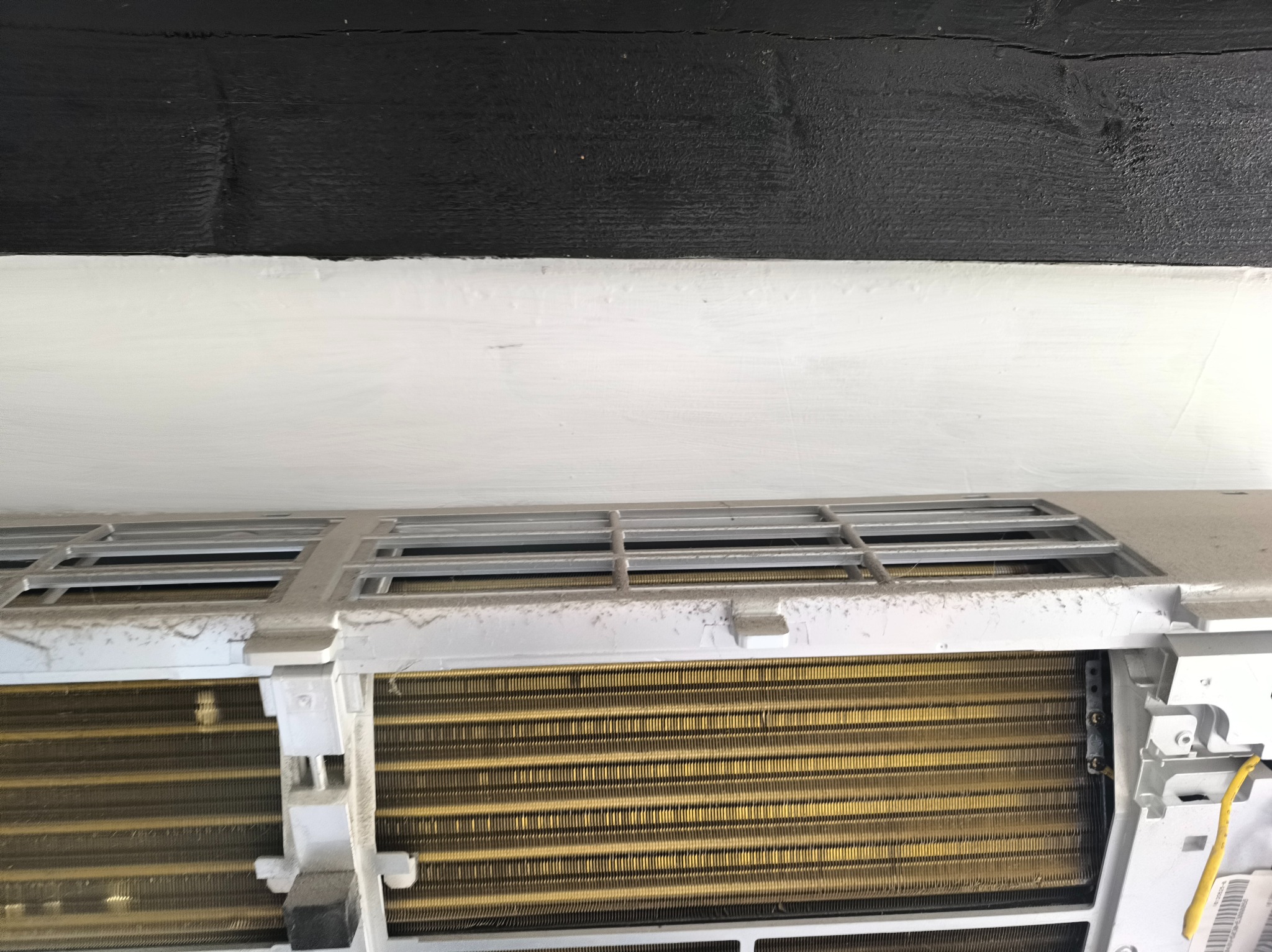 Zdemontowany, zakurzony klimatyzator typu split z odsłoniętym wymiennikiem ciepła, wiszący na białej ścianie pod czarną belką sufitową. Widoczne detale konstrukcyjne.