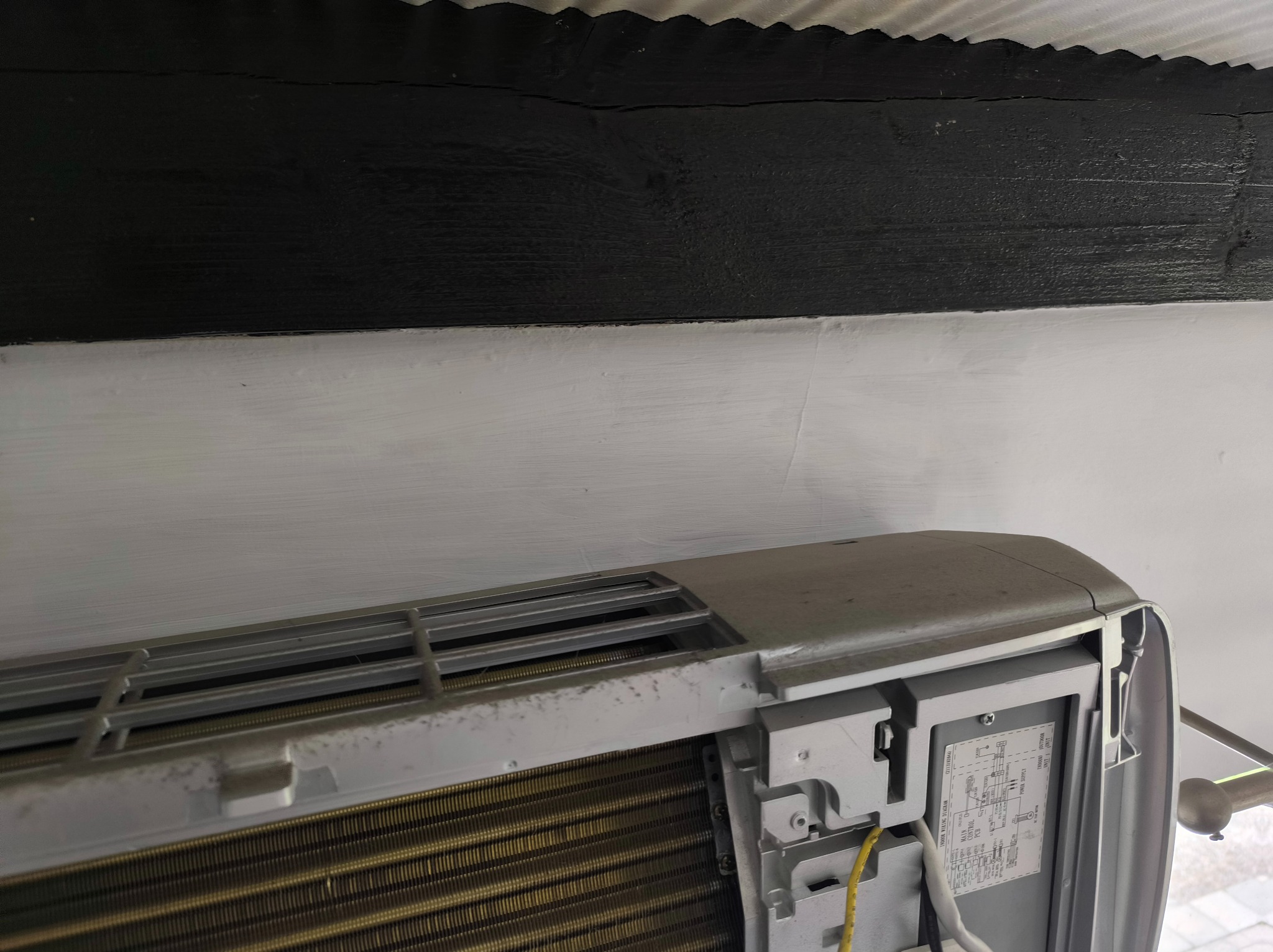 Boczny widok na klimatyzator ścienny w pomieszczeniu, częściowo widoczny czarny, drewniany element stropu. Widoczne detale konstrukcji klimatyzatora i złącza.