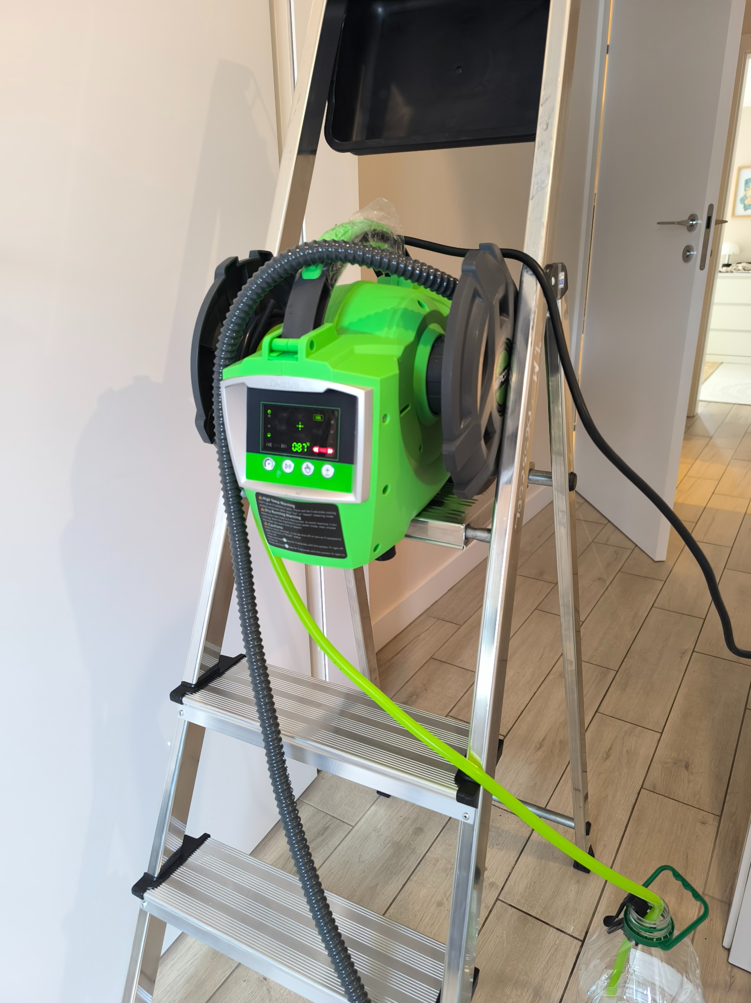 Zielone urządzenie do czyszczenia klimatyzacji, umieszczone na aluminiowej drabinie w jasnym wnętrzu. Widoczny wyświetlacz urządzenia i węże doprowadzające wodę.