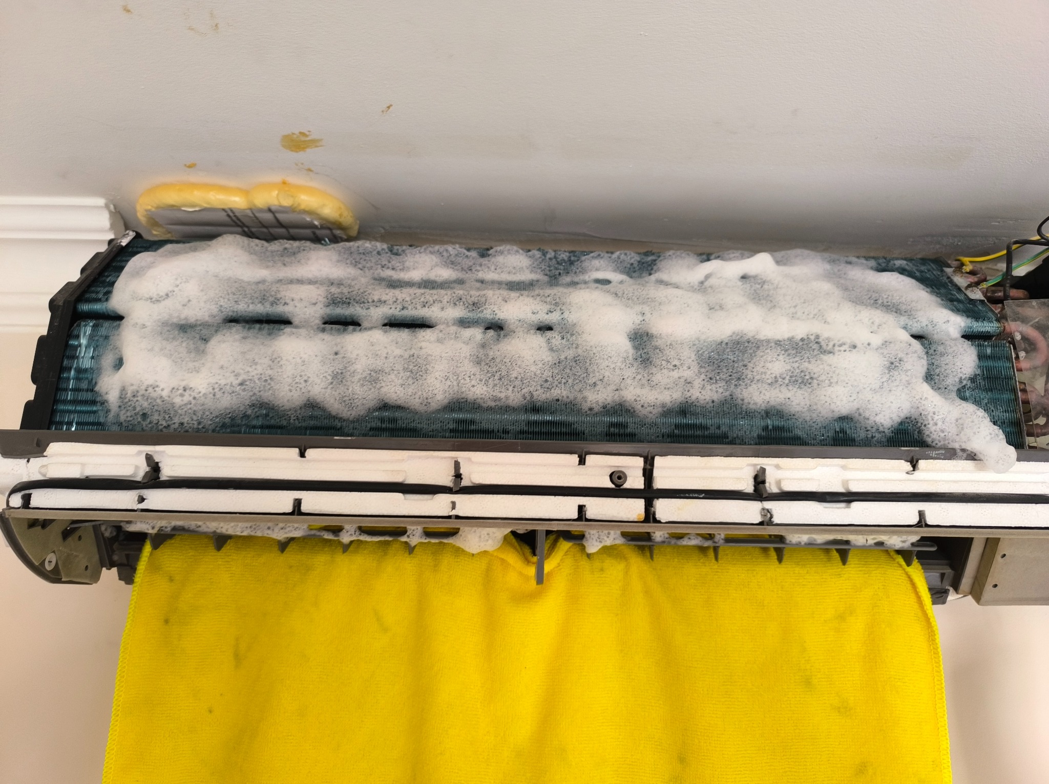 Czyszczenie klimatyzacji: widoczne piana czyszcząca na filtrach i żółta ściereczka zabezpieczająca ścianę podczas serwisu.