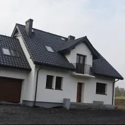 Biały dom z czarnym dachem i brązową bramą garażową, widoczny z zewnątrz, z elementami wykończeniowymi w szarym kolorze, w tle drzewa i niebo.