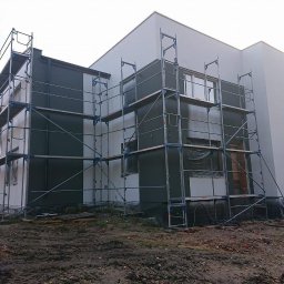 ZAKŁAD OGÓLNOBUDOWLANY DARIUSZ DĄBROWSKI - Dwukondygnacyjny dom w trakcie malowania elewacji na szaro, z rusztowaniami. Część ściany pomalowana, okna zabezpieczone folią. Teren wokół domu przygotowany do prac.