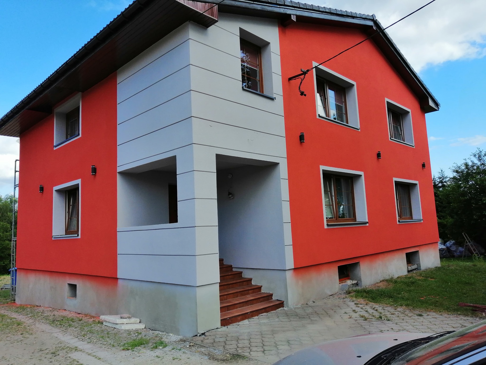 Dwukondygnacyjny dom po renowacji elewacji, z pomarańczowymi ścianami i szarymi elementami architektonicznymi, schody wejściowe wyłożone brązowymi płytkami.