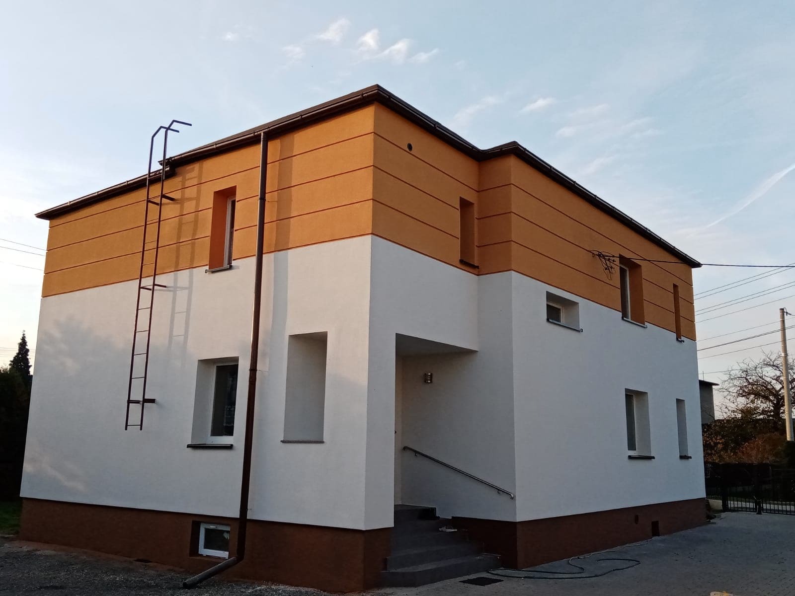 Dwukondygnacyjny dom po renowacji elewacji: biała dolna część, górna w odcieniach brązu i beżu. Widoczna drabina przymocowana do ściany.