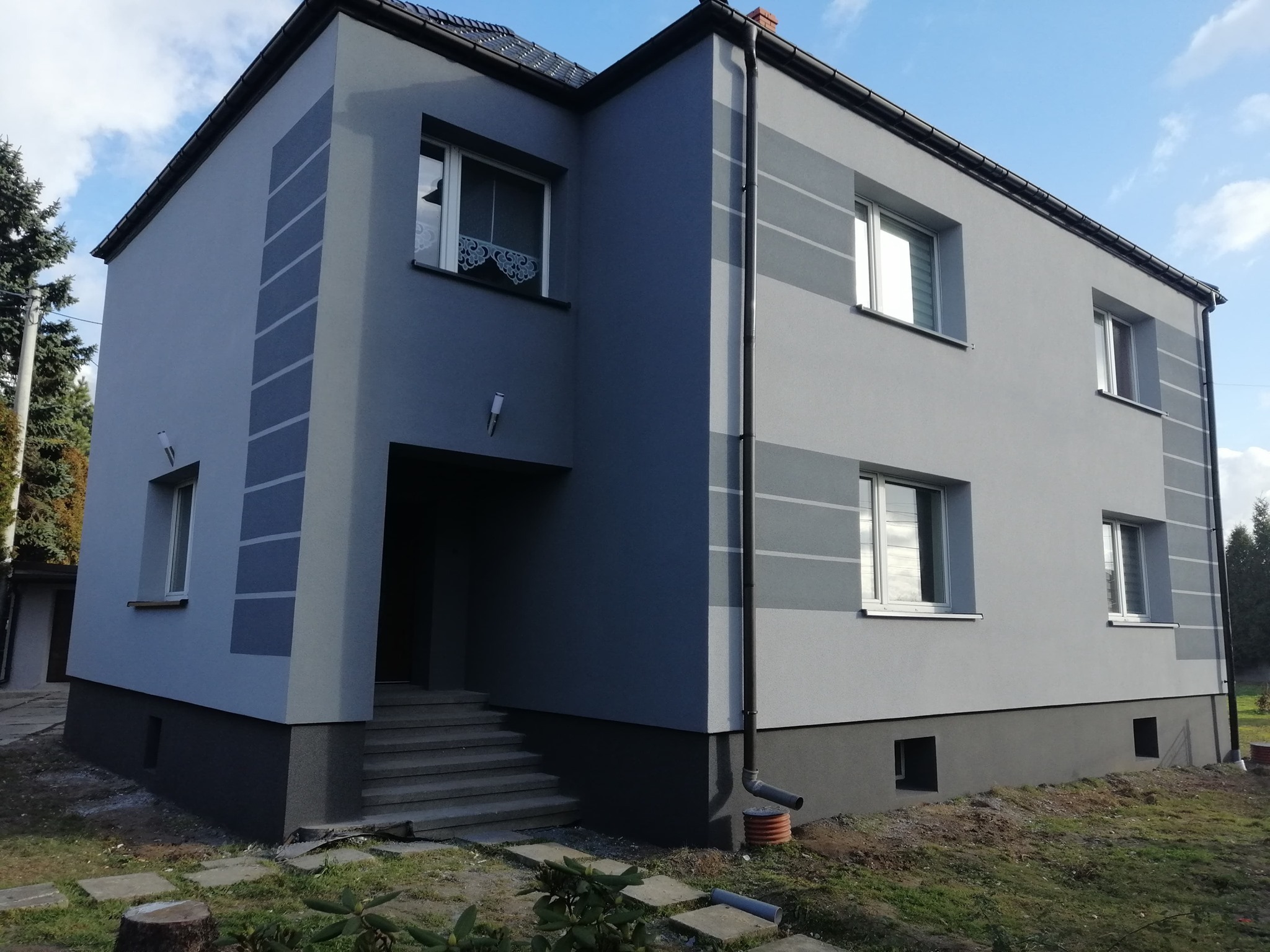 Dwukondygnacyjny dom w odcieniach szarości z ciemnoszarą podmurówką i dekoracyjnymi pasami na elewacji, widok z zewnątrz w słoneczny dzień.