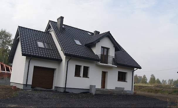 Biały dom z czarnym dachem i brązową bramą garażową, widoczny z zewnątrz, z elementami wykończeniowymi w szarym kolorze, w tle drzewa i niebo.