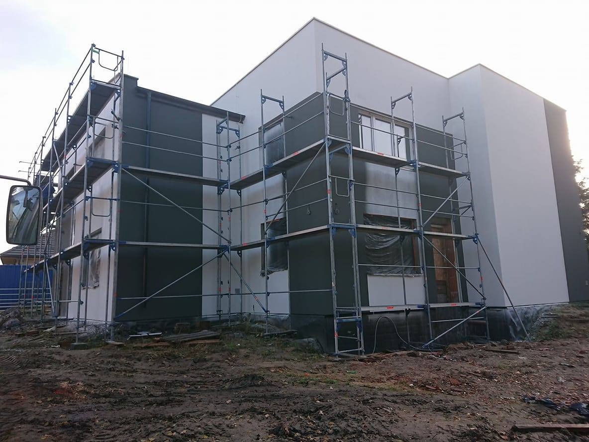 Dwukondygnacyjny dom w trakcie malowania elewacji na szaro, z rusztowaniami. Część ściany pomalowana, okna zabezpieczone folią. Teren wokół domu przygotowany do prac.