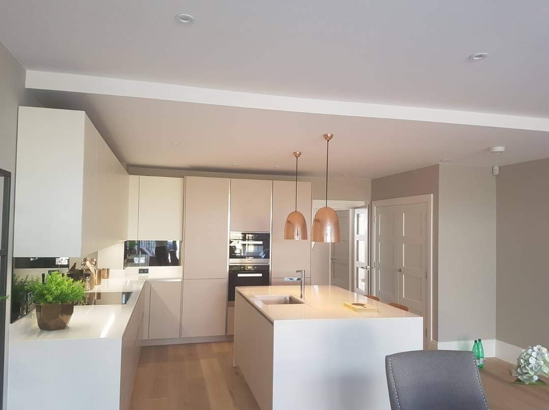 Nowoczesna, jasna kuchnia z wyspą i wiszącymi lampami miedzianymi. Minimalistyczne fronty szafek, drewniana podłoga i rośliny doniczkowe dodają elegancji wnętrzu.