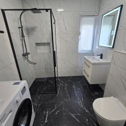 Remont pod klucz - Wykończona łazienka z czarno-białą marmurową okładziną, kabiną prysznicową typu walk-in z czarną ramą, wiszącą toaletą i pralką Samsung.