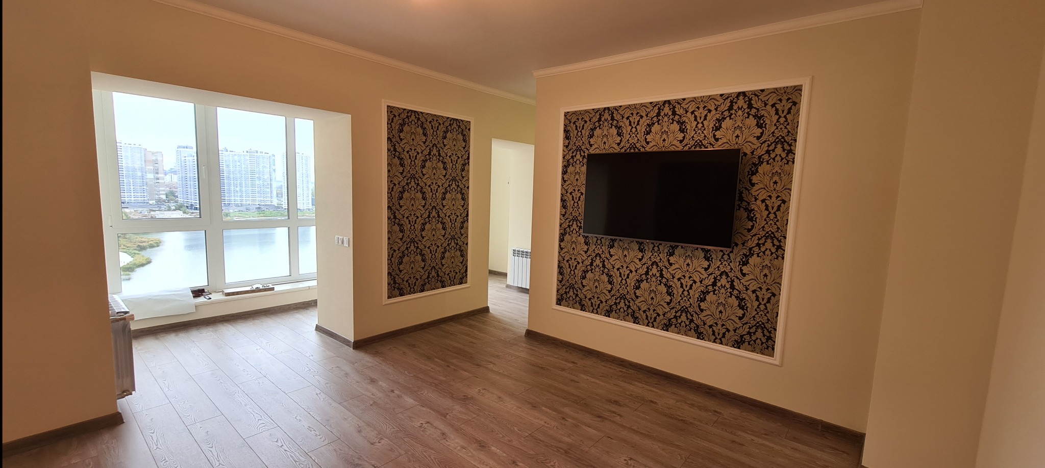 Salon po remoncie z panelami podłogowymi, oknem z widokiem na miasto, dekoracyjną tapetą w ramie i zawieszonym telewizorem. Jasne ściany i listwy przypodłogowe.