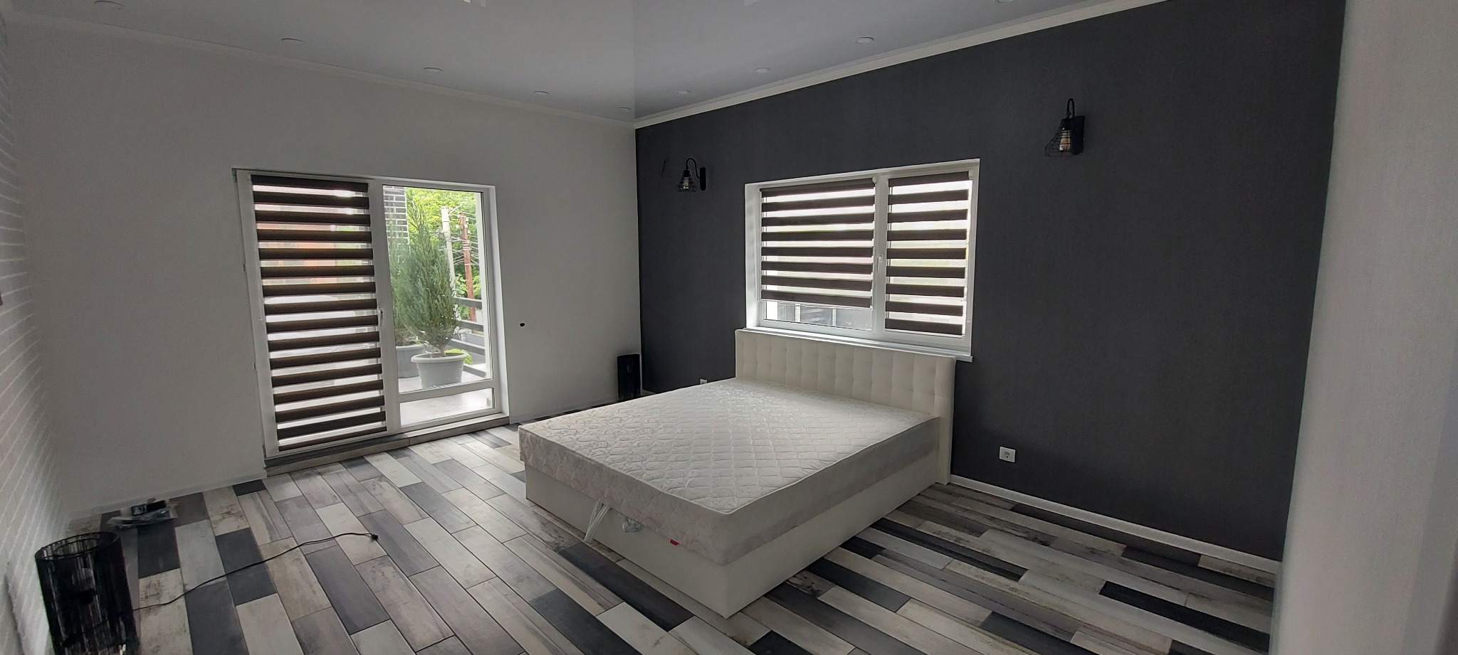 Elegancka sypialnia z podwójnym łóżkiem, szarą ścianą z lampami i drewnopodobną podłogą. Widok na balkon z roślinami przez okno z żaluzjami.