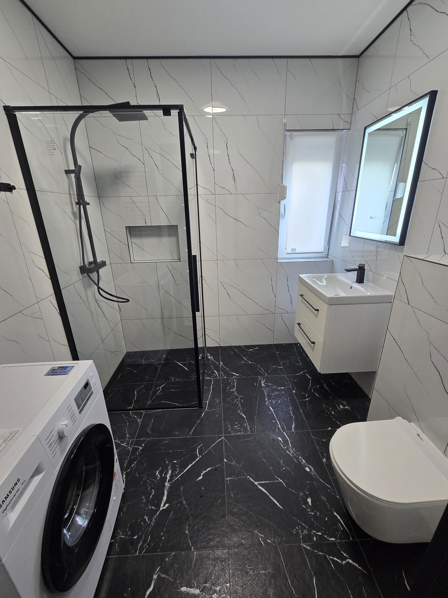 Wykończona łazienka z czarno-białą marmurową okładziną, kabiną prysznicową typu walk-in z czarną ramą, wiszącą toaletą i pralką Samsung.