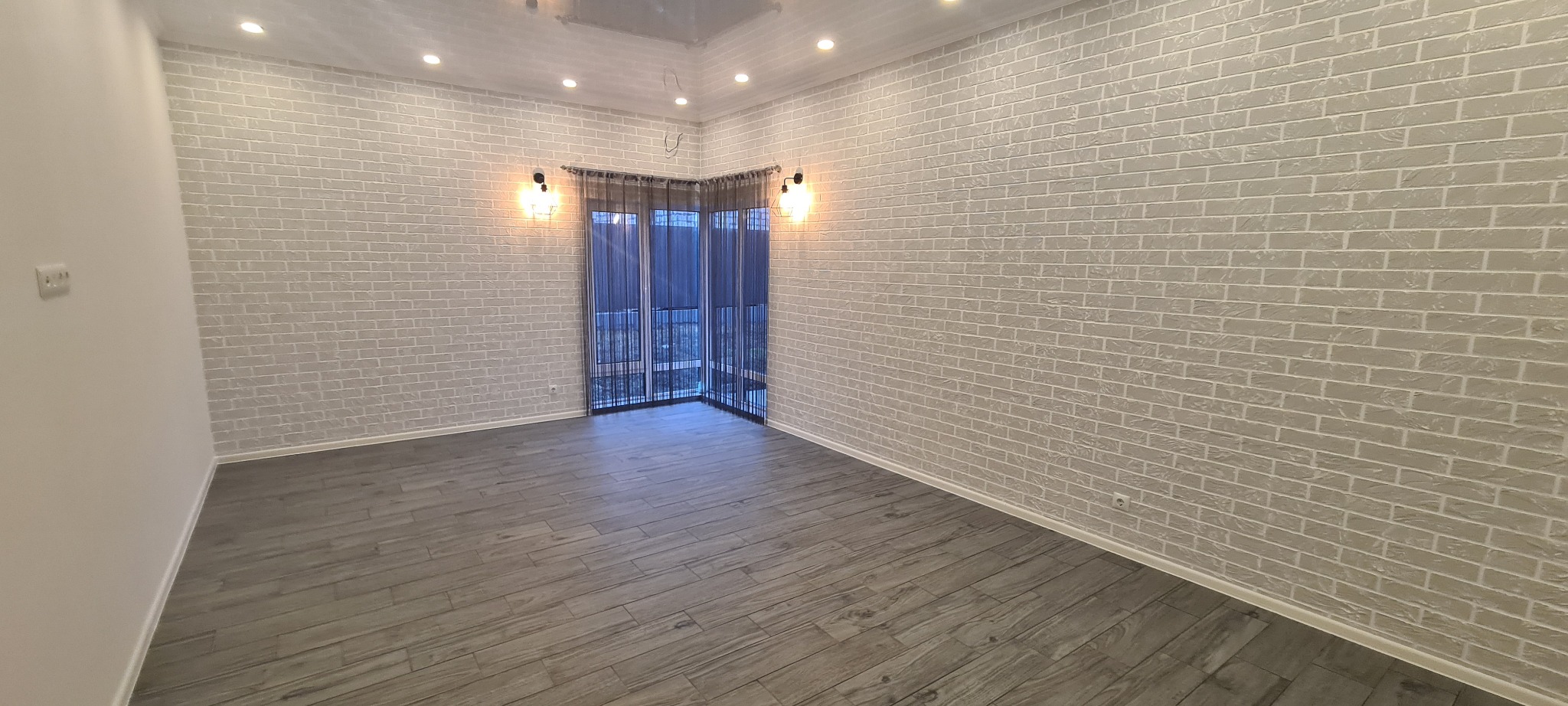 Pokój po remoncie: ściany z efektem cegły w odcieniach szarości, drewnopodobna podłoga, okno z ciemnymi zasłonami i lampy na ścianach. Nowoczesne wykończenie wnętrza.