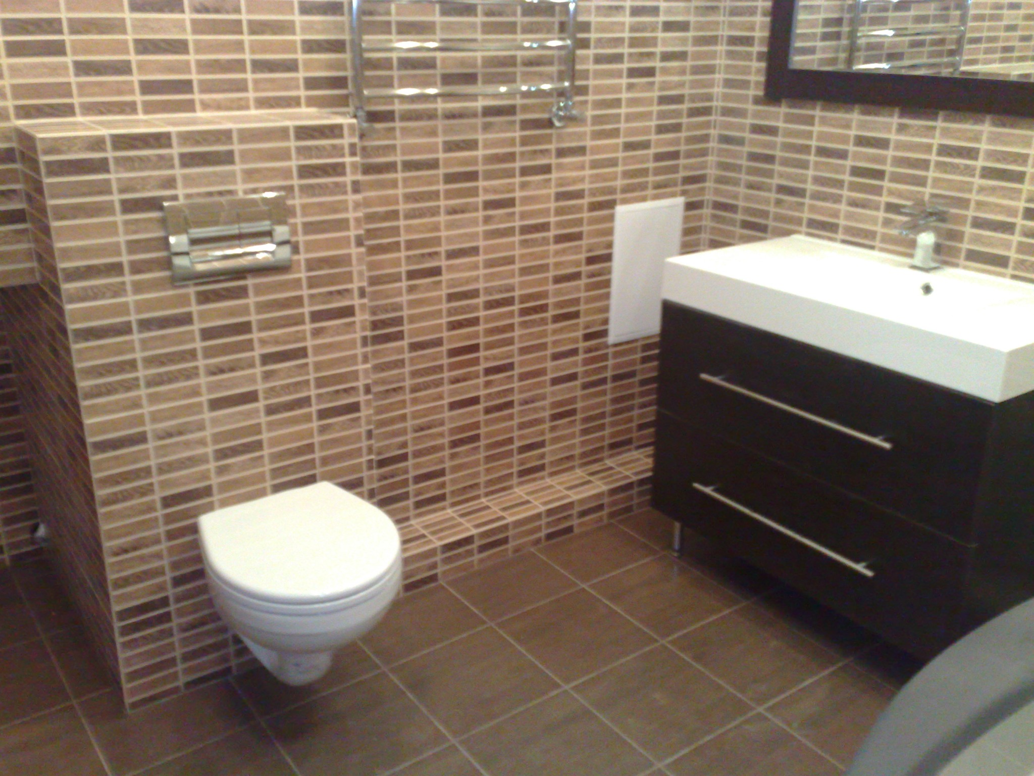 Wykończona łazienka z brązowymi płytkami mozaikowymi, wiszącą toaletą i szafką z umywalką. Grzejnik drabinkowy i lustro w tle.