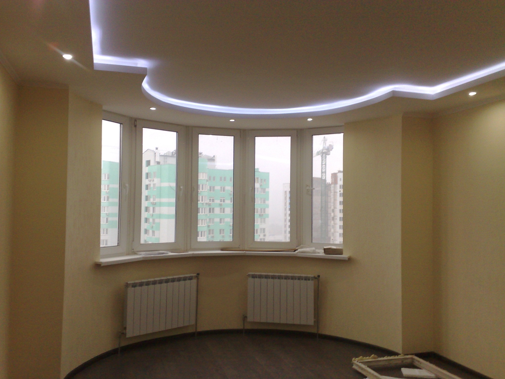 Nowoczesny salon z oknem wykuszowym i podwieszanym sufitem z oświetleniem LED. Widok na budynek w tle. Dwa grzejniki pod oknem. Podłoga w ciemnym odcieniu brązu.