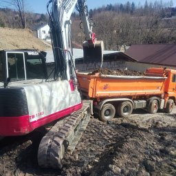 DAW-POL Sylwester Dawiec - Koparka Takeuchi TB 1140 nabiera ziemię do pomarańczowej wywrotki MAN na placu budowy. W tle zabudowa i drzewa. Słoneczny dzień.