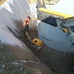 DAW-POL Sylwester Dawiec - Praca przy wykopie: mężczyzna ubija ziemię zagęszczarką skoczną. Wykop zabezpieczony folią i geowłókniną, widoczny fragment koparki. Ujęcie z góry.