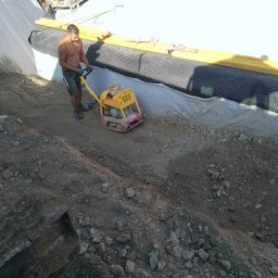 DAW-POL Sylwester Dawiec - Pracownik ubija ziemię zagęszczarką skoczkową w wąskim wykopie z zabezpieczonymi ścianami, widoczne materiały budowlane w tle. Praca w upalny dzień.