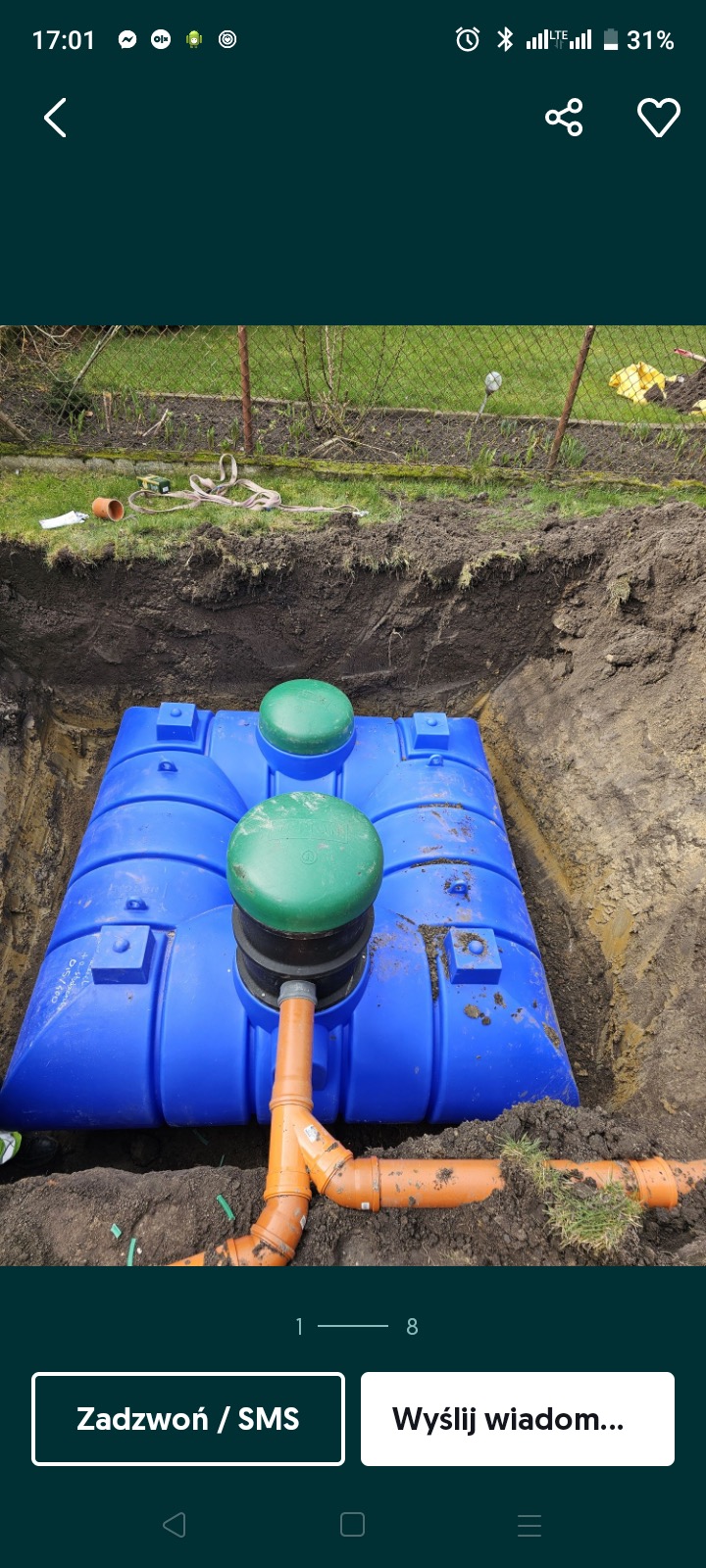 Niebieski zbiornik z zielonymi pokrywami w wykopie, widoczne rury kanalizacyjne. Trawa i siatka ogrodzeniowa w tle. Ujęcie z góry ukazuje etap instalacji.