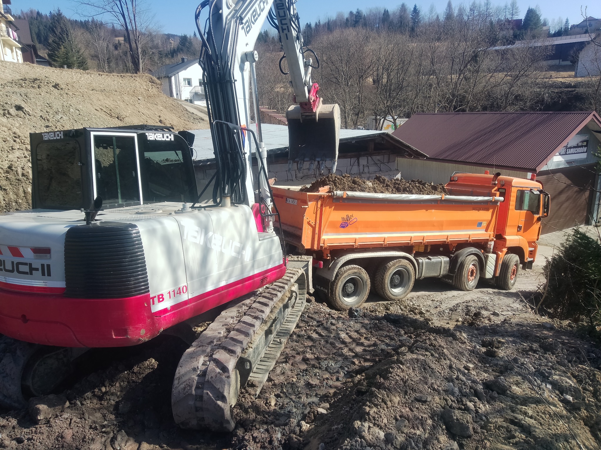 Koparka Takeuchi TB 1140 nabiera ziemię do pomarańczowej wywrotki MAN na placu budowy. W tle zabudowa i drzewa. Słoneczny dzień.