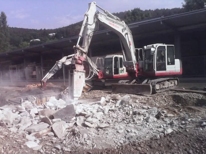 Biała koparka z pneumatycznym młotem kruszy skały na placu budowy. W tle widoczny zadaszony obszar i zalesione wzgórze.