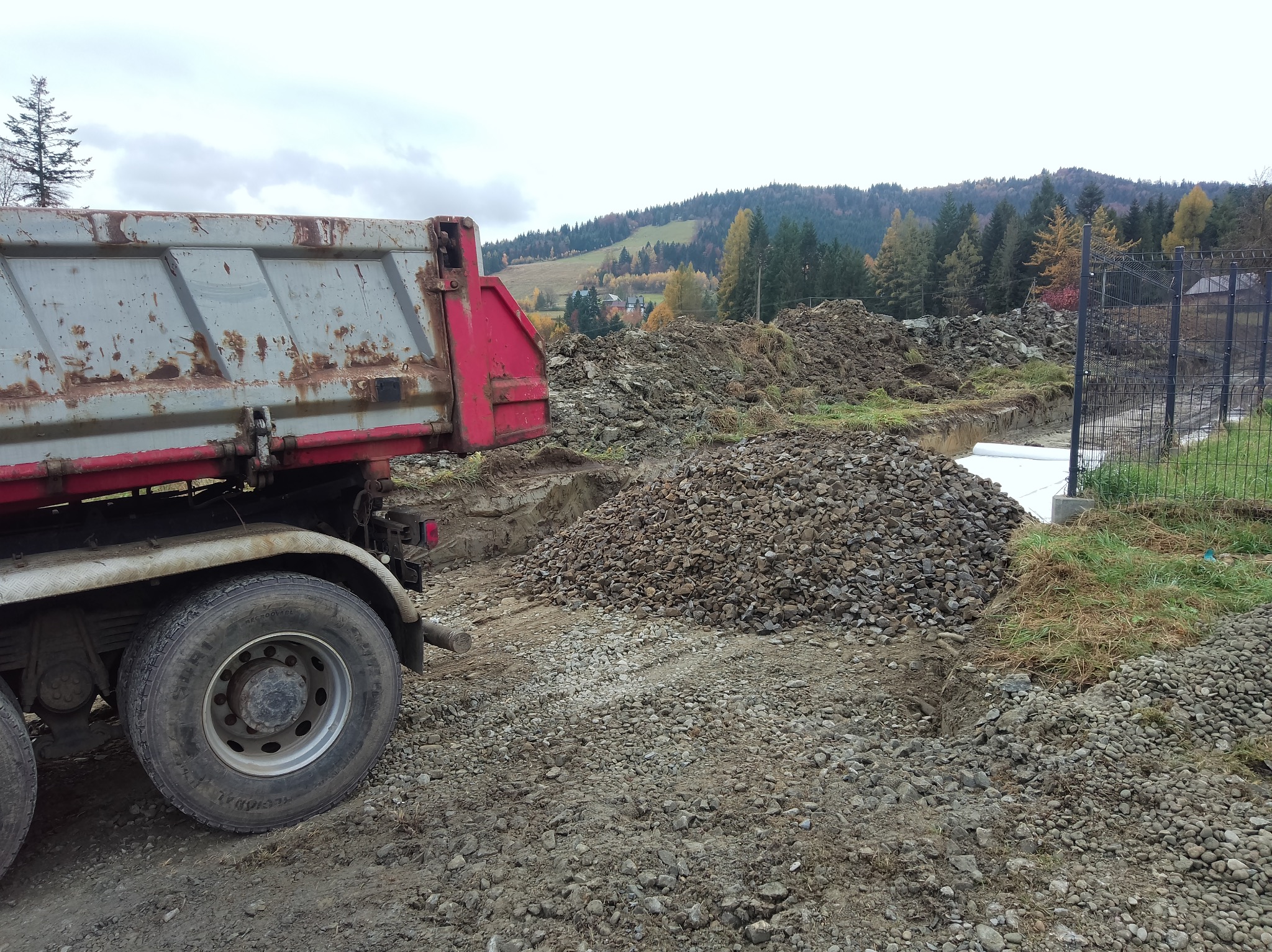 Ciężarówka wywrotka z korozją, obok hałda gruzu i wykopu pod fundamenty, w tle wzgórza z lasem w jesiennej scenerii.