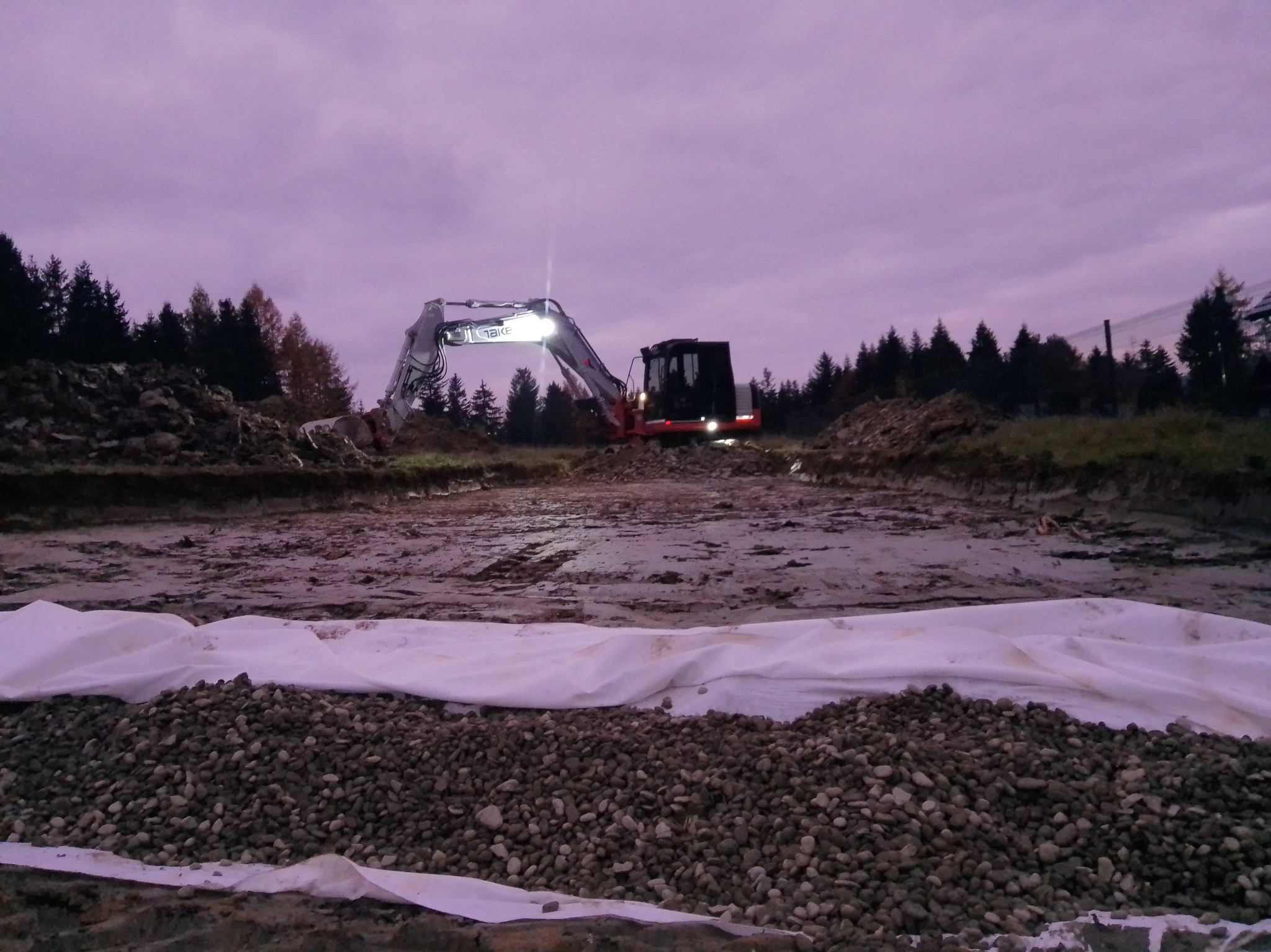 Wieczorny widok na teren budowy: koparka z włączonymi światłami, kamienie na geowłókninie i przygotowany teren pod drenaż w otoczeniu drzew.