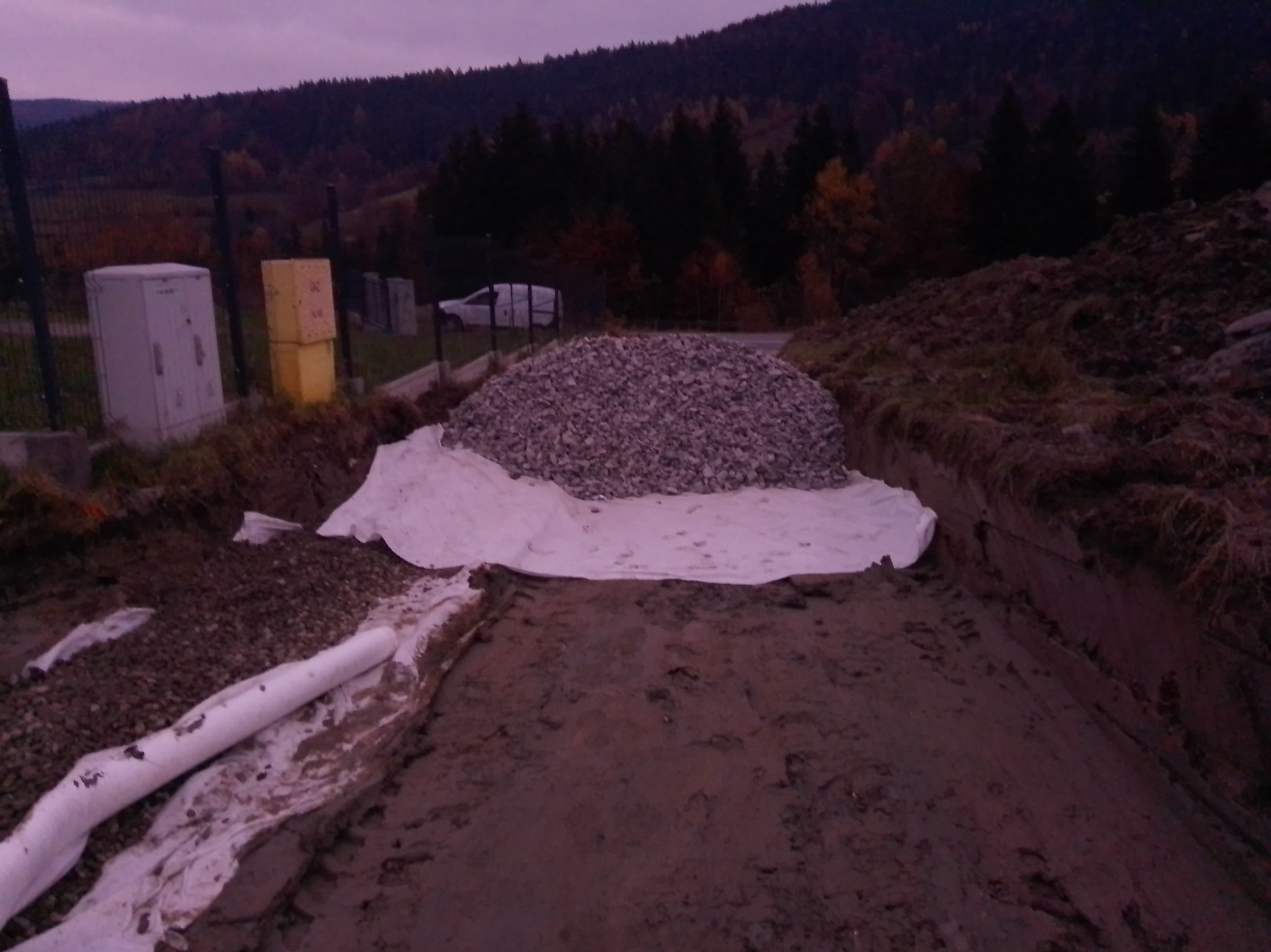 Kopuła żwiru na białej geowłókninie w wykopie, w tle las i zabudowania. Widoczne rury drenażowe i wykopany grunt. Ujęcie z poziomu gruntu, pochmurne niebo.