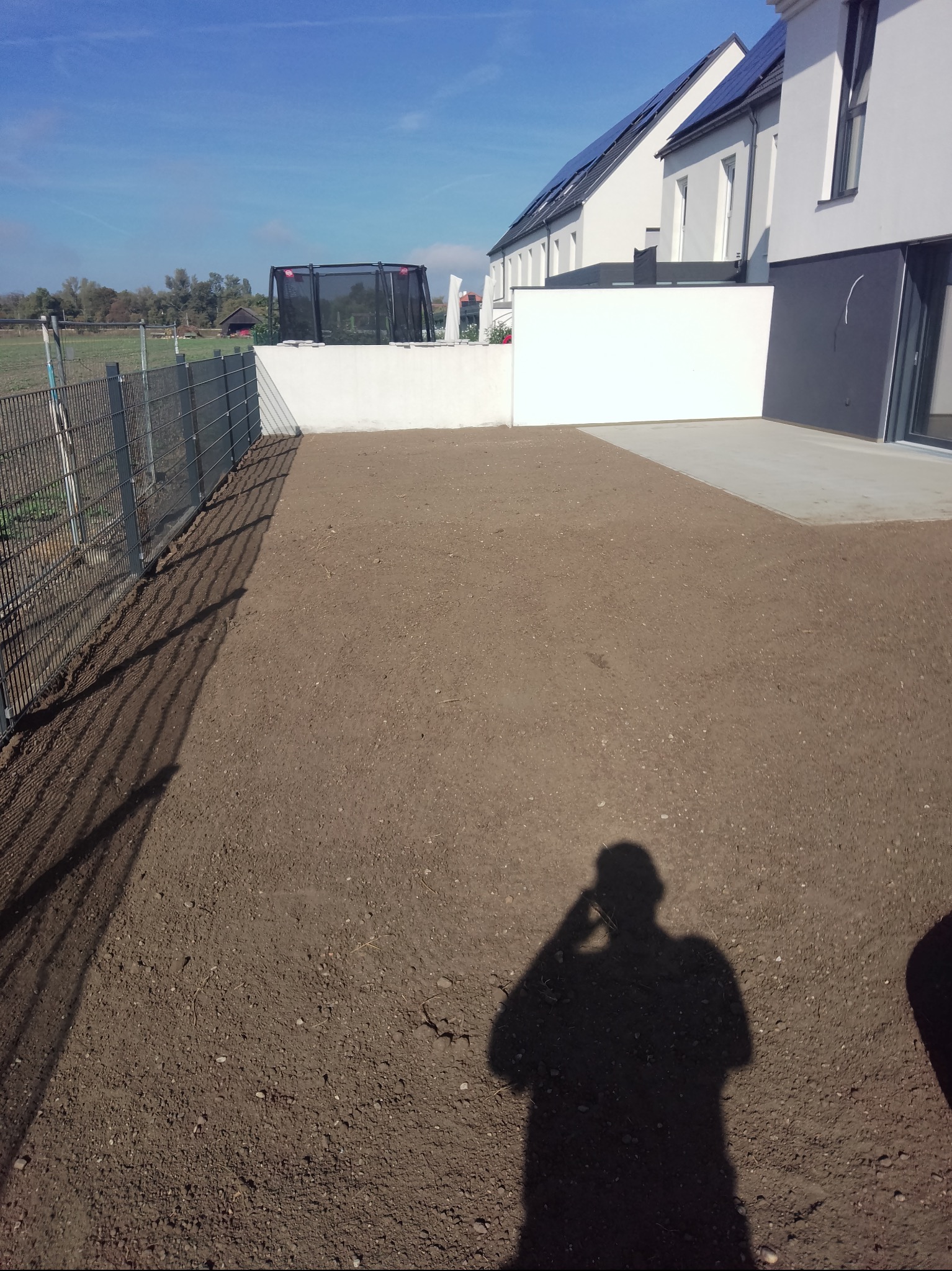 Przygotowany teren pod trawnik przy nowoczesnym domu z panelami słonecznymi. Widoczny cień osoby robiącej zdjęcie. Ogrodzenie z siatki po lewej.