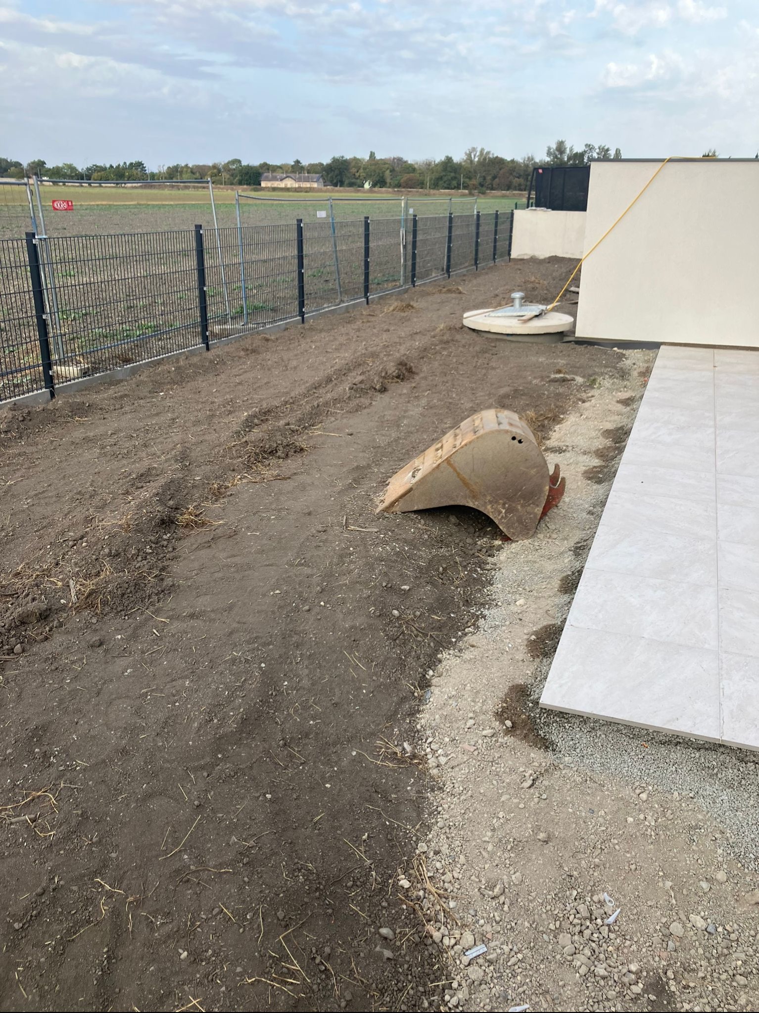 Teren po niwelacji, widoczny fragment ogrodzenia panelowego, studzienka kanalizacyjna i łyżka koparki obok ułożonych płytek. Ślady opon na ziemi.