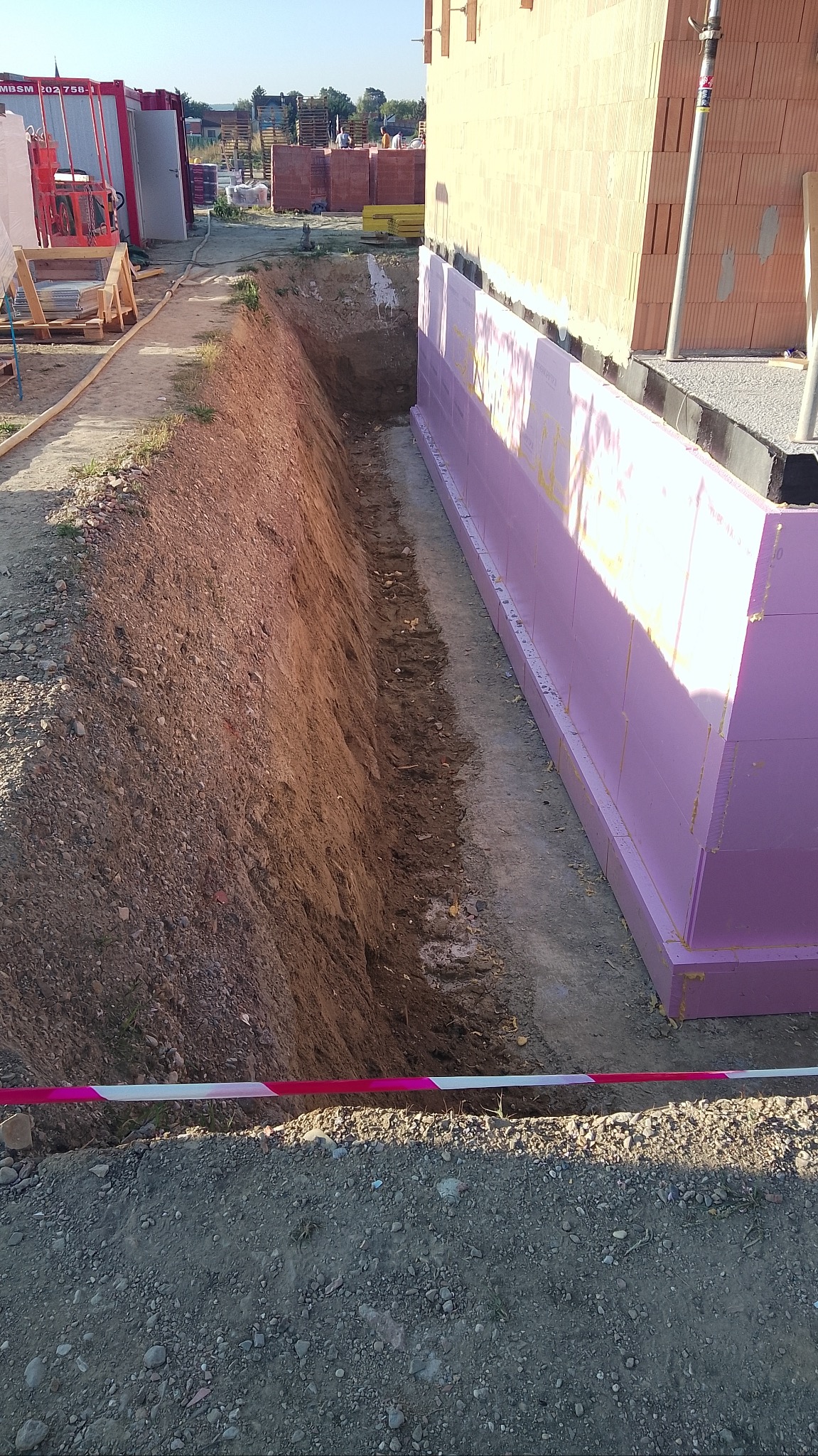 Wykop pod fundamenty budynku z izolacją termiczną z różowego styropianu. W tle widoczne materiały budowlane i kontener. Ujęcie z poziomu gruntu.