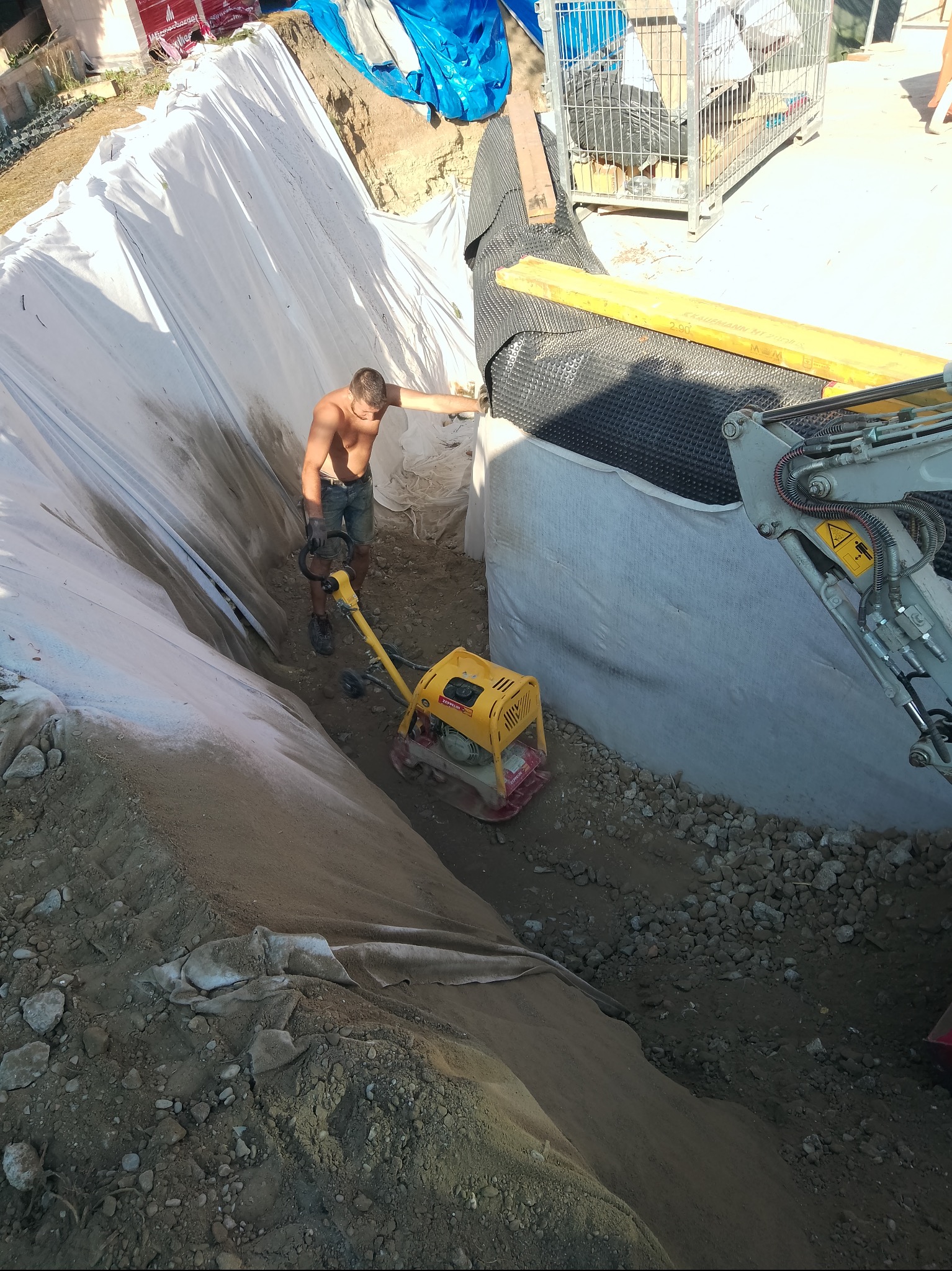 Praca przy wykopie: mężczyzna ubija ziemię zagęszczarką skoczną. Wykop zabezpieczony folią i geowłókniną, widoczny fragment koparki. Ujęcie z góry.