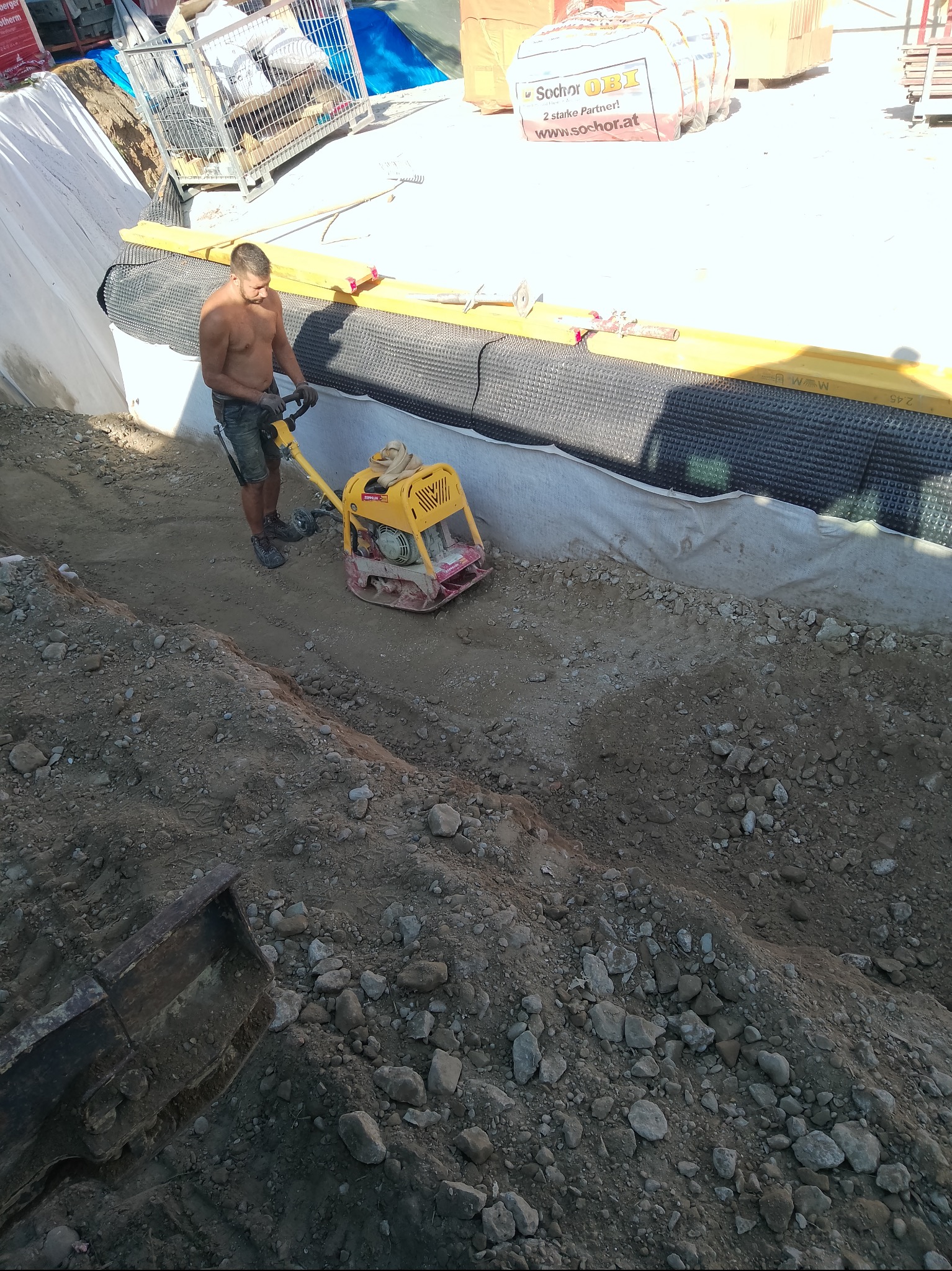 Pracownik ubija ziemię zagęszczarką skoczkową w wąskim wykopie z zabezpieczonymi ścianami, widoczne materiały budowlane w tle. Praca w upalny dzień.