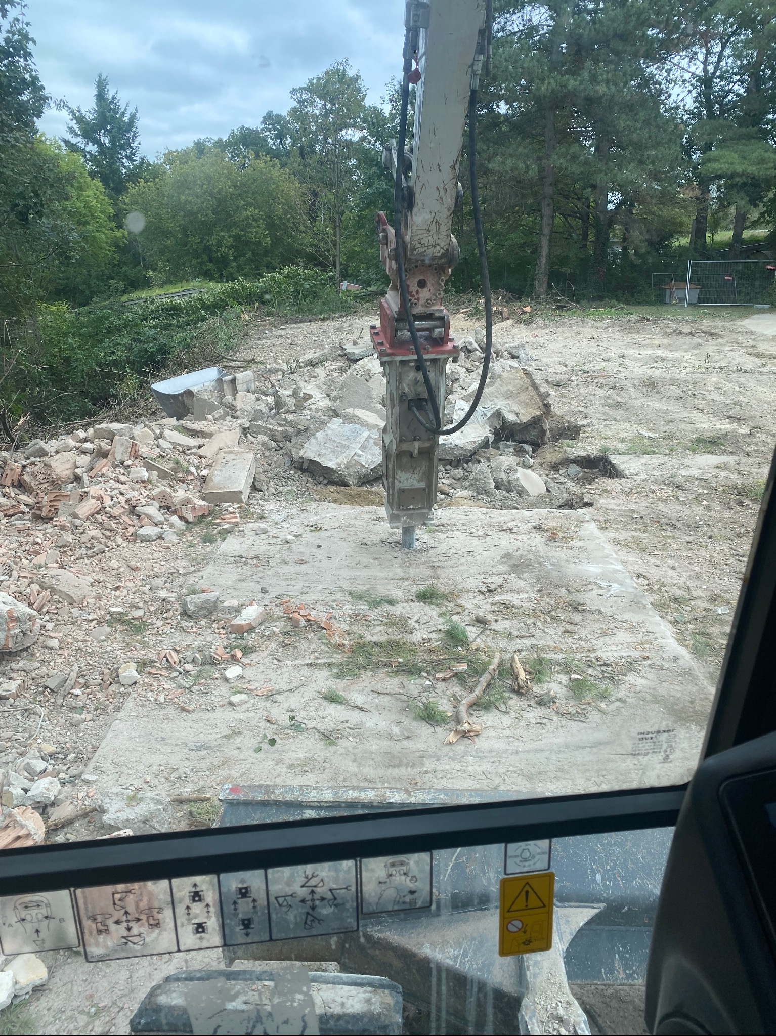 Widok z kabiny na młot hydrauliczny kruszący betonową płytę. W tle gruz i zieleń. Maszyna w trakcie pracy, teren po rozbiórce.