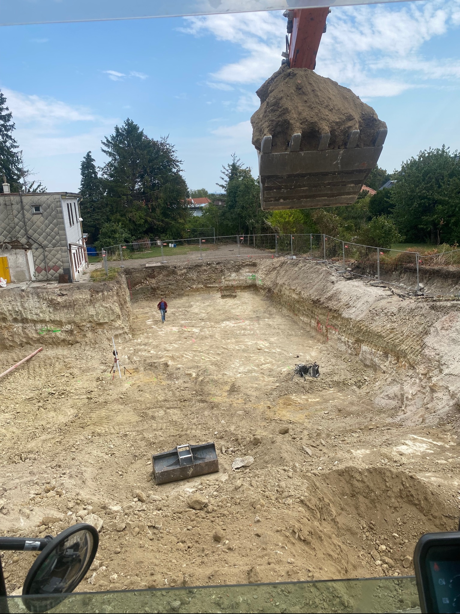 Głęboki wykop pod fundamenty z pracującą koparką, widok z góry. W tle budynek i zieleń. Na dnie wykopu stoi człowiek i sprzęt geodezyjny.