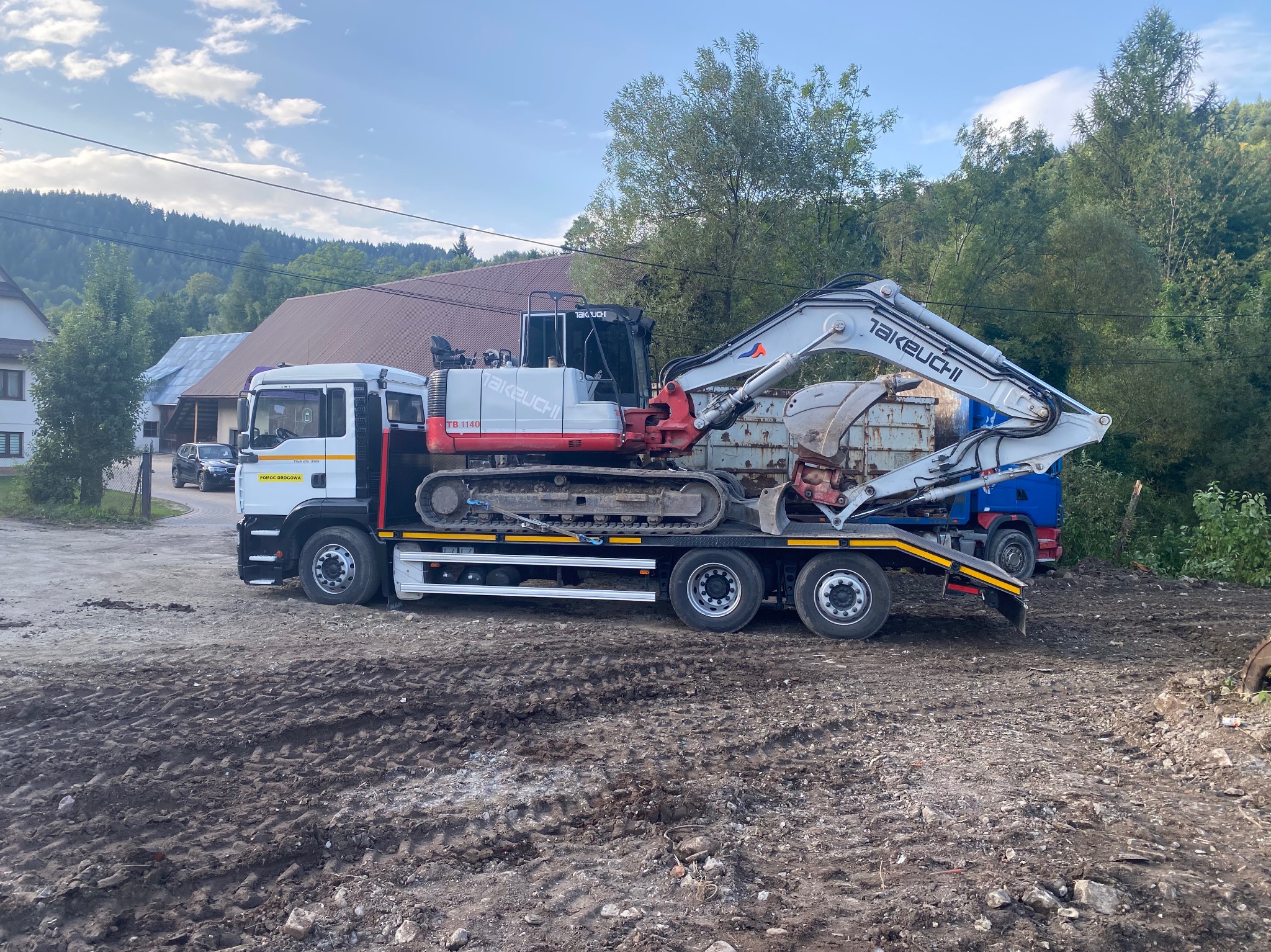 Ciężarówka z niskopodwoziową naczepą przewozi koparkę Takeuchi TB 1140 na tle wiejskiego krajobrazu z domami i drzewami. Widoczne ślady opon na ziemi.