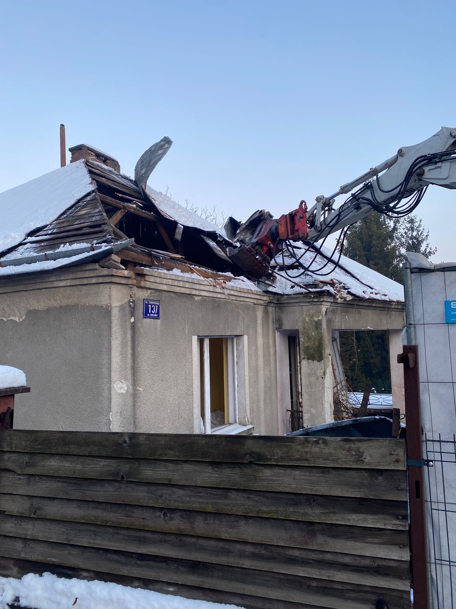 Rozbiórka domu jednorodzinnego w zimowej scenerii. Częściowo zdemontowany dach, widoczny gruz i maszyna budowlana z mechanicznym ramieniem. Tabliczka z adresem na ścianie.