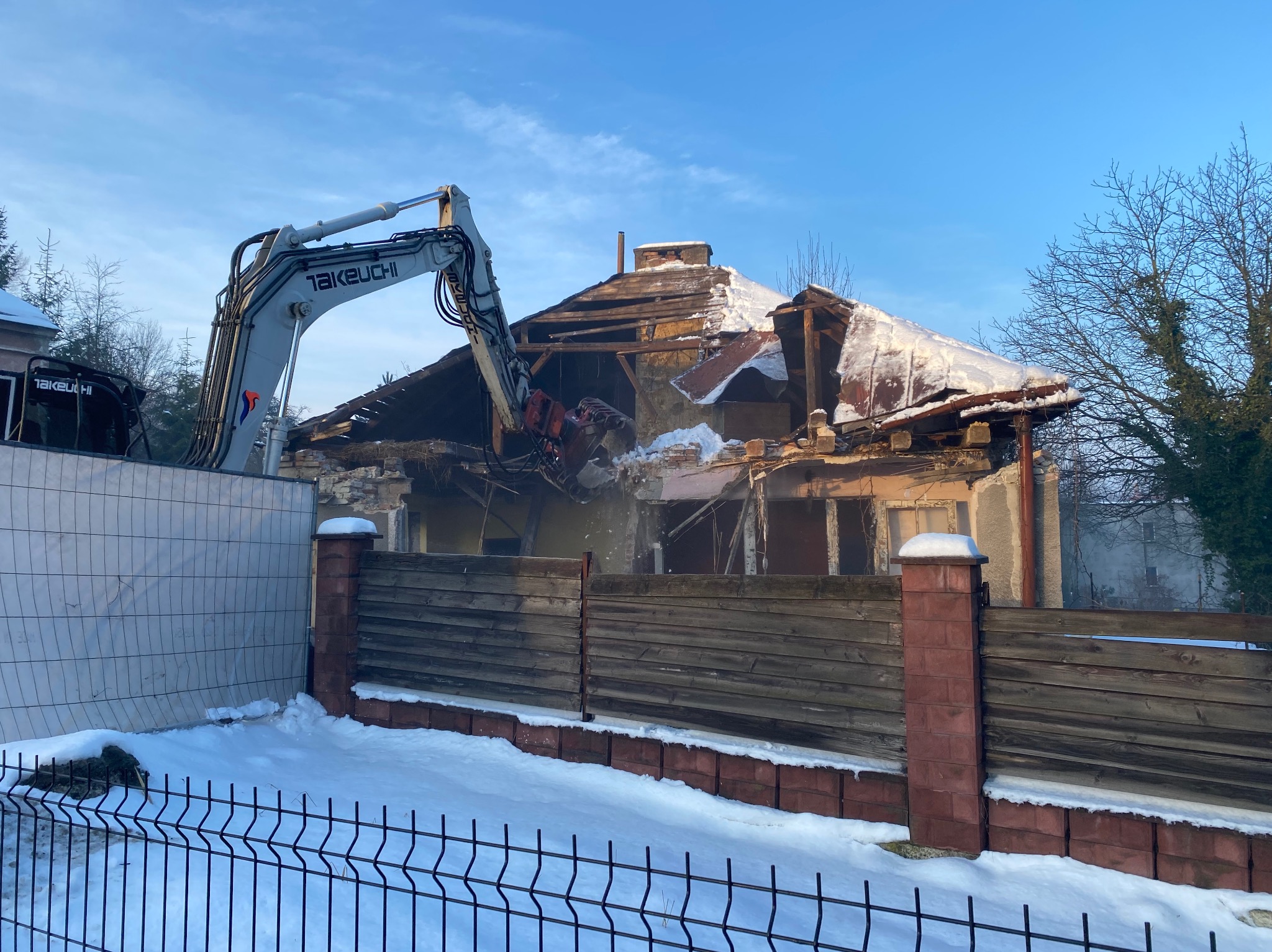 Demolka domu jednorodzinnego w zimowej scenerii. Koparka Takeuchi w trakcie rozbiórki dachu, widoczne fragmenty ścian i gruzu. Częściowo ośnieżony teren i ogrodzenie.