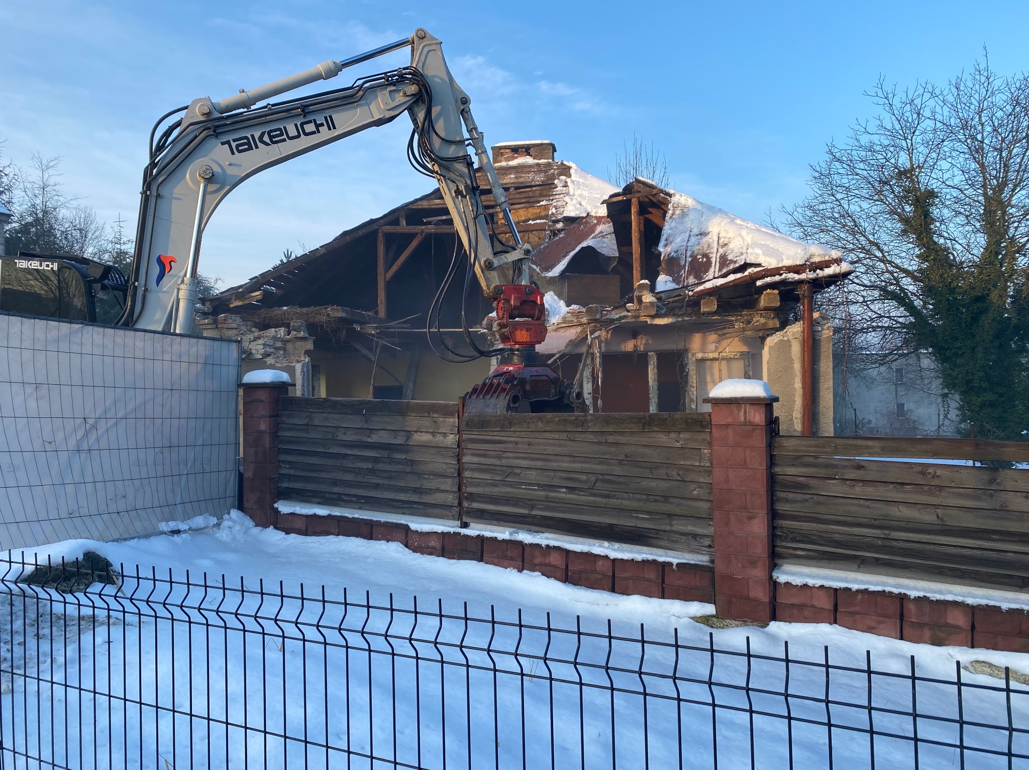 Rozbiórka domu jednorodzinnego w zimowej scenerii. Koparka Takeuchi z hydraulicznymi nożycami kruszy resztki dachu. W tle drzewa bez liści i błękitne niebo.