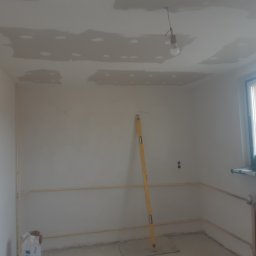 Osoba prywatna - Pokój w trakcie prac wykończeniowych: szpachlowane fragmenty sufitu, poziomica oparta o ścianę, widoczne instalacje. Przy oknie narzędzia i grzejnik.